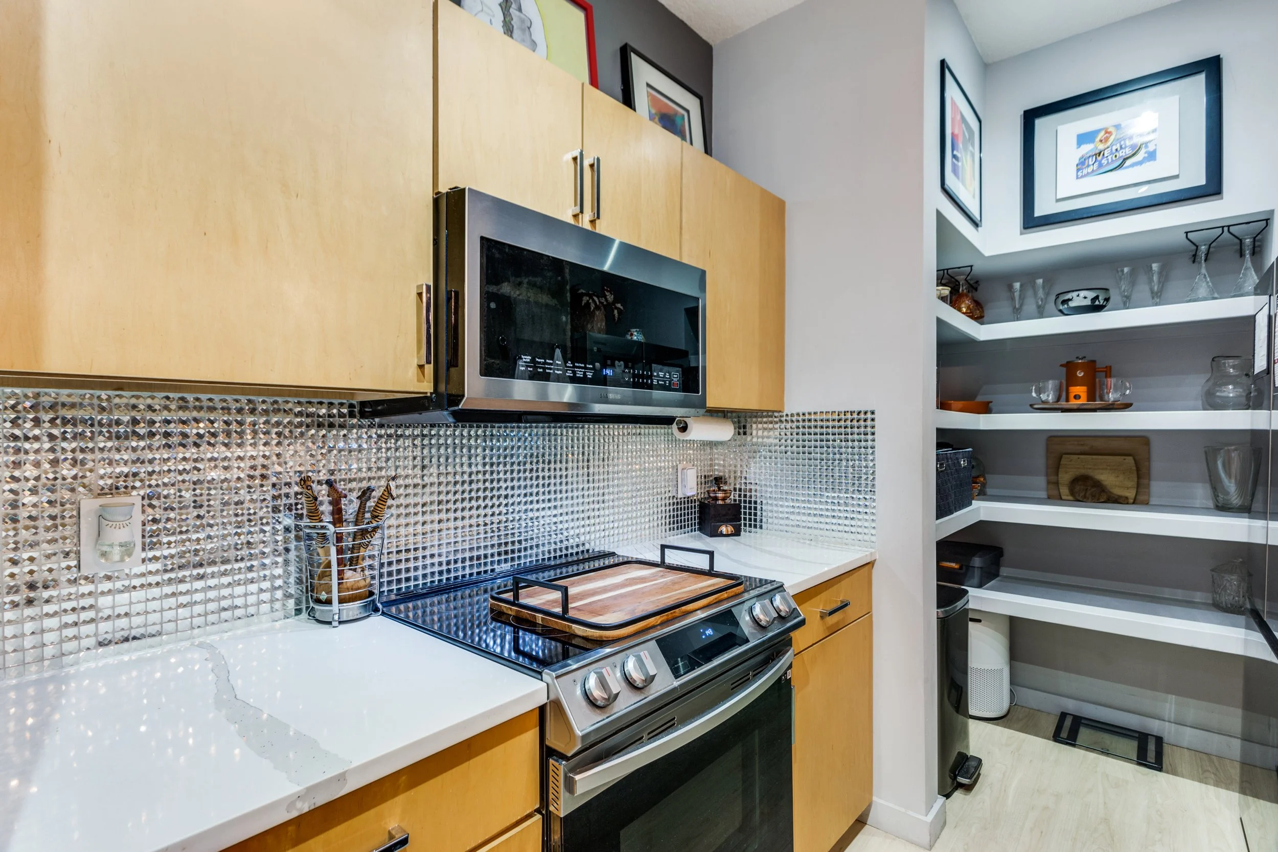 Kitchen with wooden cabinets, a microwave above the stove, a cutting board on the stove, and a pantry with glassware and decorative items on shelves.