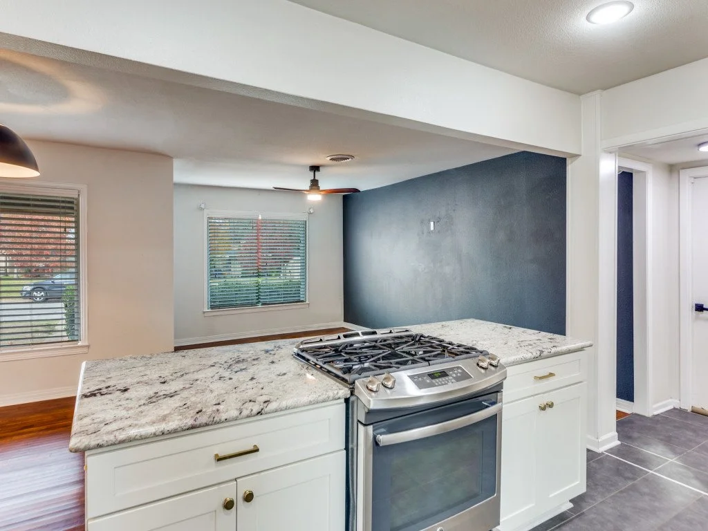 View of a modern kitchen with white cabinets and a granite countertop, stainless steel stove, dark accent wall, and adjacent living area with windows and blinds.