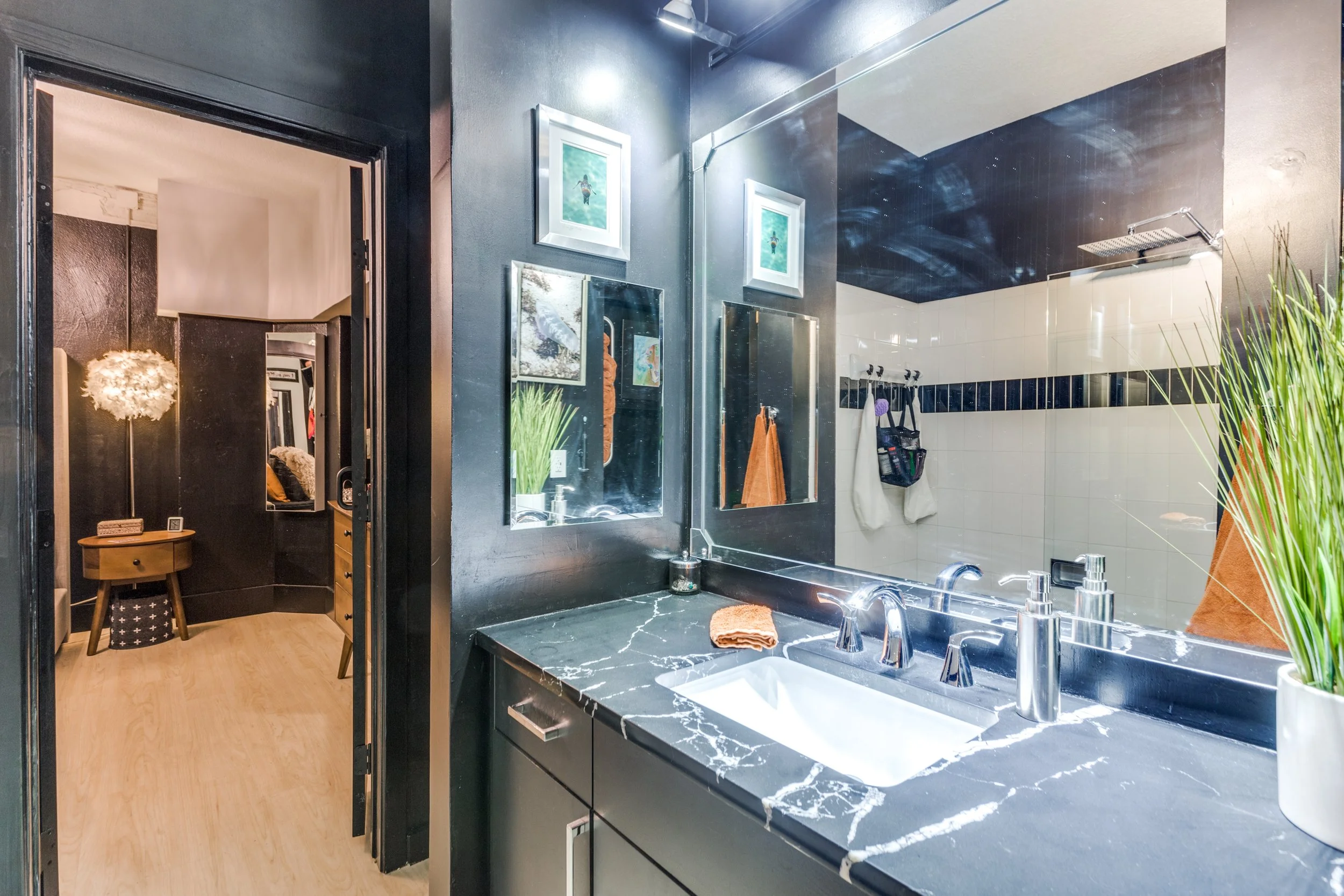 Modern bathroom with black and white marble vanity, large mirror, and glass shower with white tiles; decor includes framed pictures, potted plant, and orange towels.