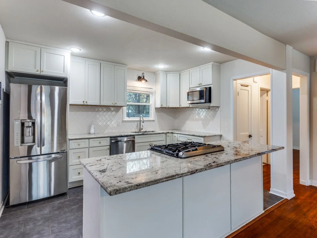 Modern kitchen with white cabinets, stainless steel refrigerator, microwave, and dishwasher, granite countertop island, gas stovetop, illuminated by ceiling lights and a small window.