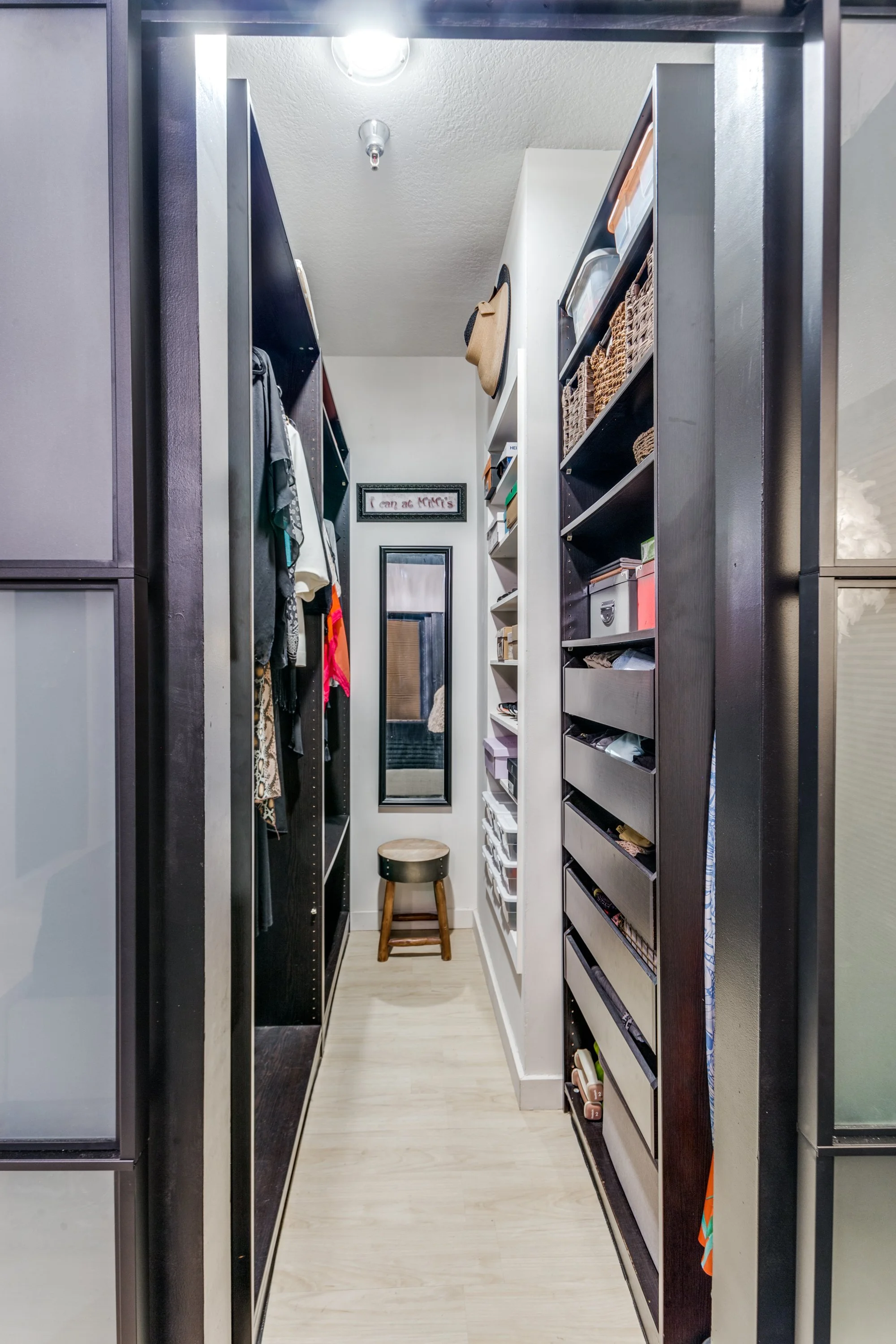 A narrow walk-in closet with black and white shelving units filled with clothing, boxes, and accessories, a small stool at the end, a full-length mirror on the back wall, and framed wall art above the mirror.