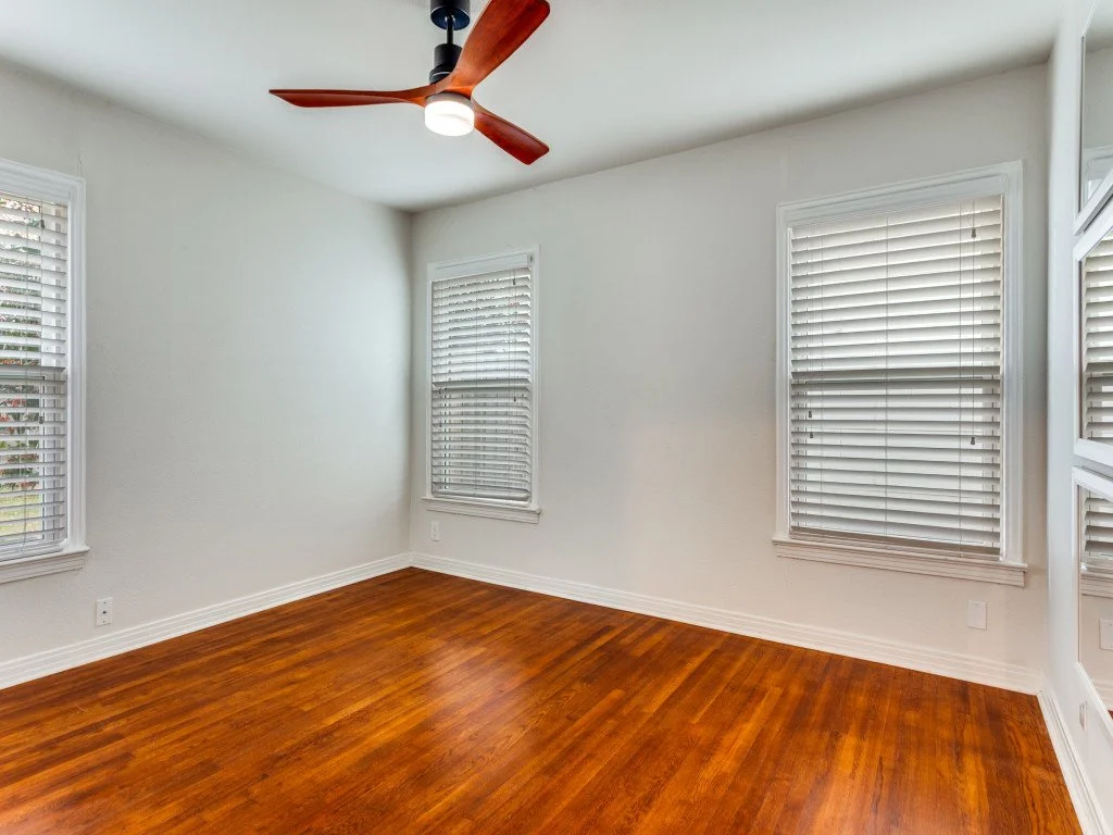 Empty room with white walls, four windows with white blinds, brown hardwood floors, and a ceiling fan with wooden blades and a modern light fixture.