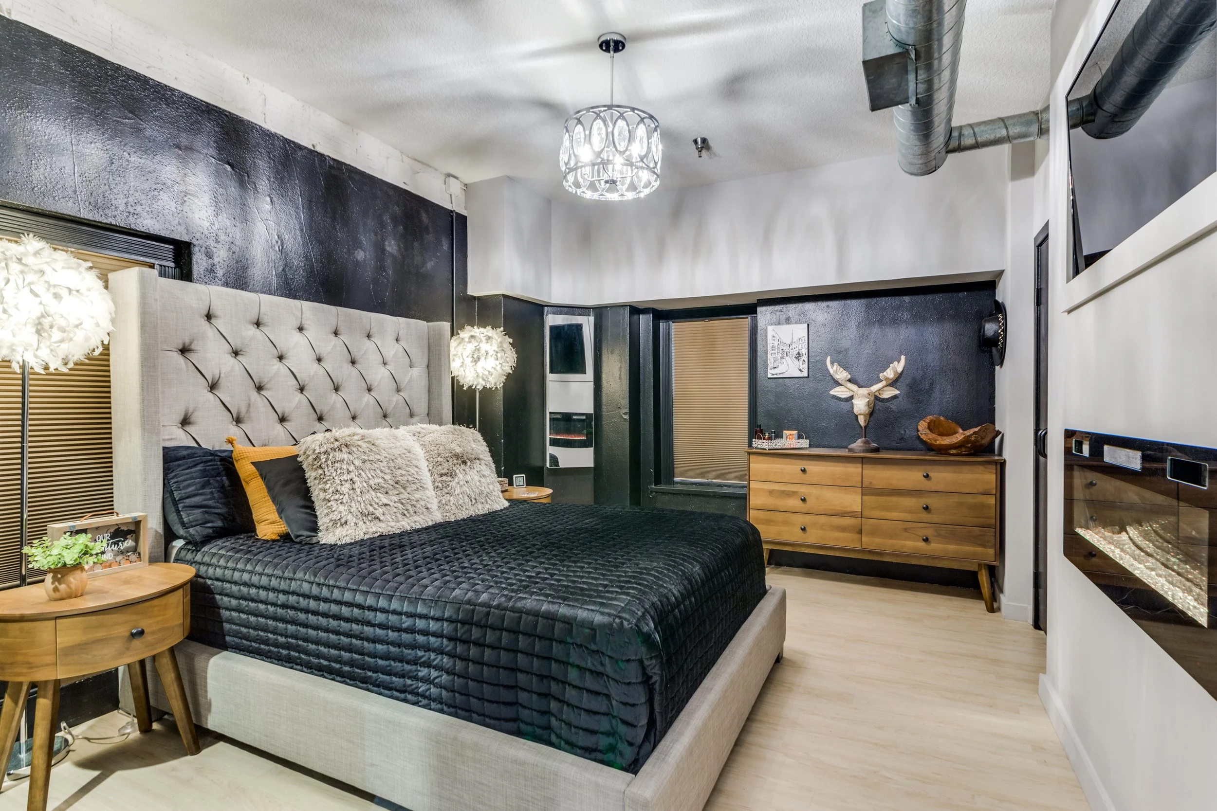 Modern bedroom with a large beige tufted headboard, black bedding, and fluffy pillows. Wooden nightstand with a small plant and picture frame. Two wall-mounted lamps with fuzzy white shades. A wooden dresser with decorative sculpture and a window wit