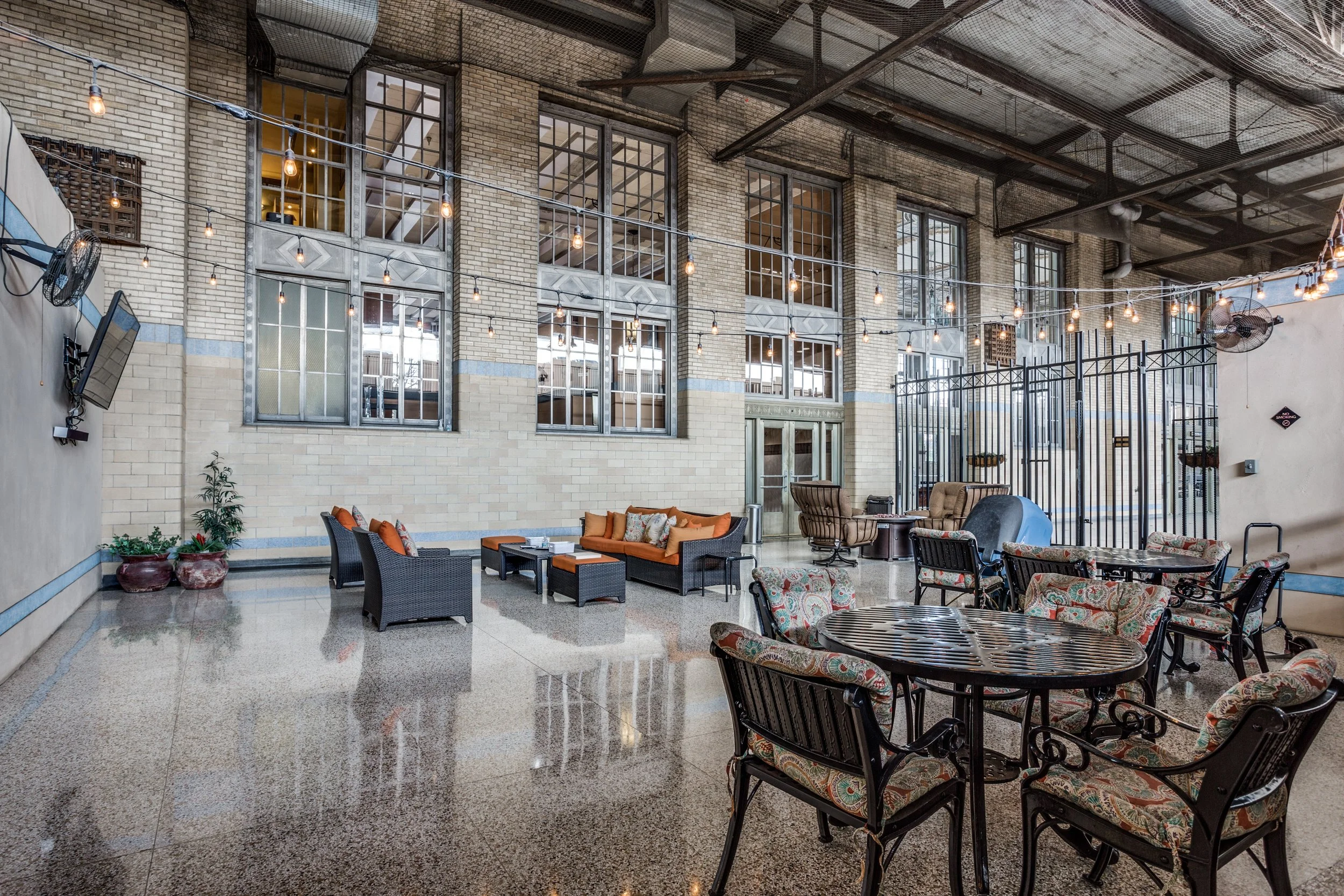 Indoor lounge or social area with wicker and upholstered chairs and tables, decorative string lights, brick walls with large metal-framed windows, potted plants, and industrial-style ceiling with metal beams.