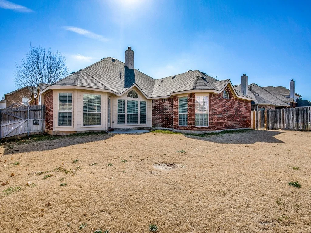 Backyard view of a brick and beige house with large windows and a chimney, surrounded by a wooden fence on a sunny day.