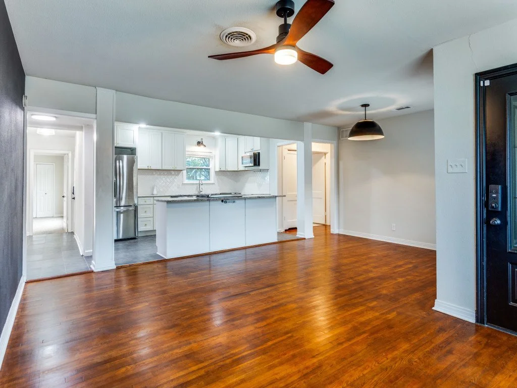 Empty living room with hardwood floors, white walls, ceiling fan, and open kitchen with white cabinets, stainless steel appliances, and a small window.
