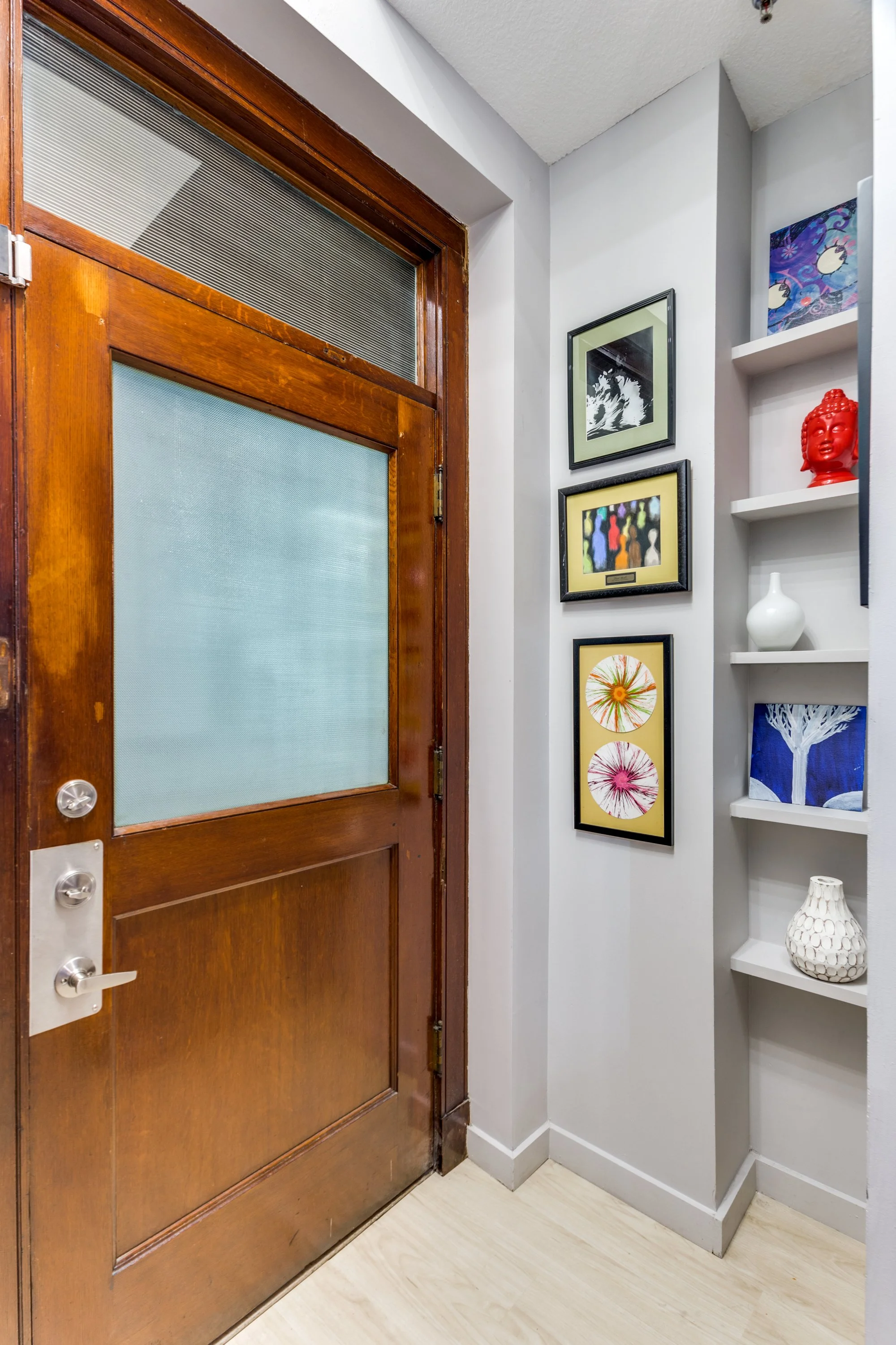 Interior view of a doorway with a wooden door, next to a white wall decorated with three framed artworks, and built-in shelves with decorative objects including vases and sculptures.