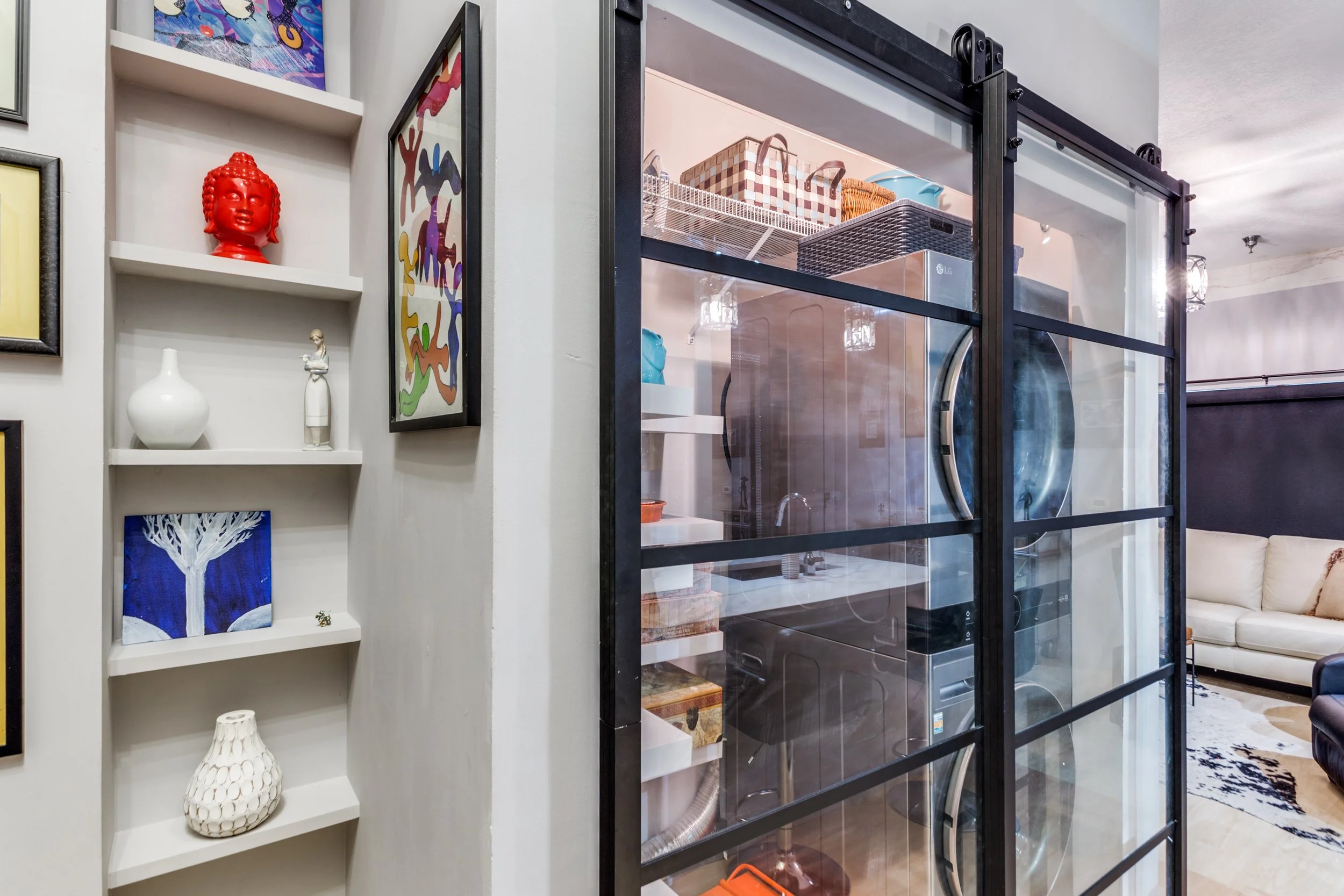 A laundry room with a glass door, laundry machines, and multiple shelves with storage baskets. Next to it, a white wall with built-in shelves holds decorative vases, artwork, and a red Buddha head sculpture.