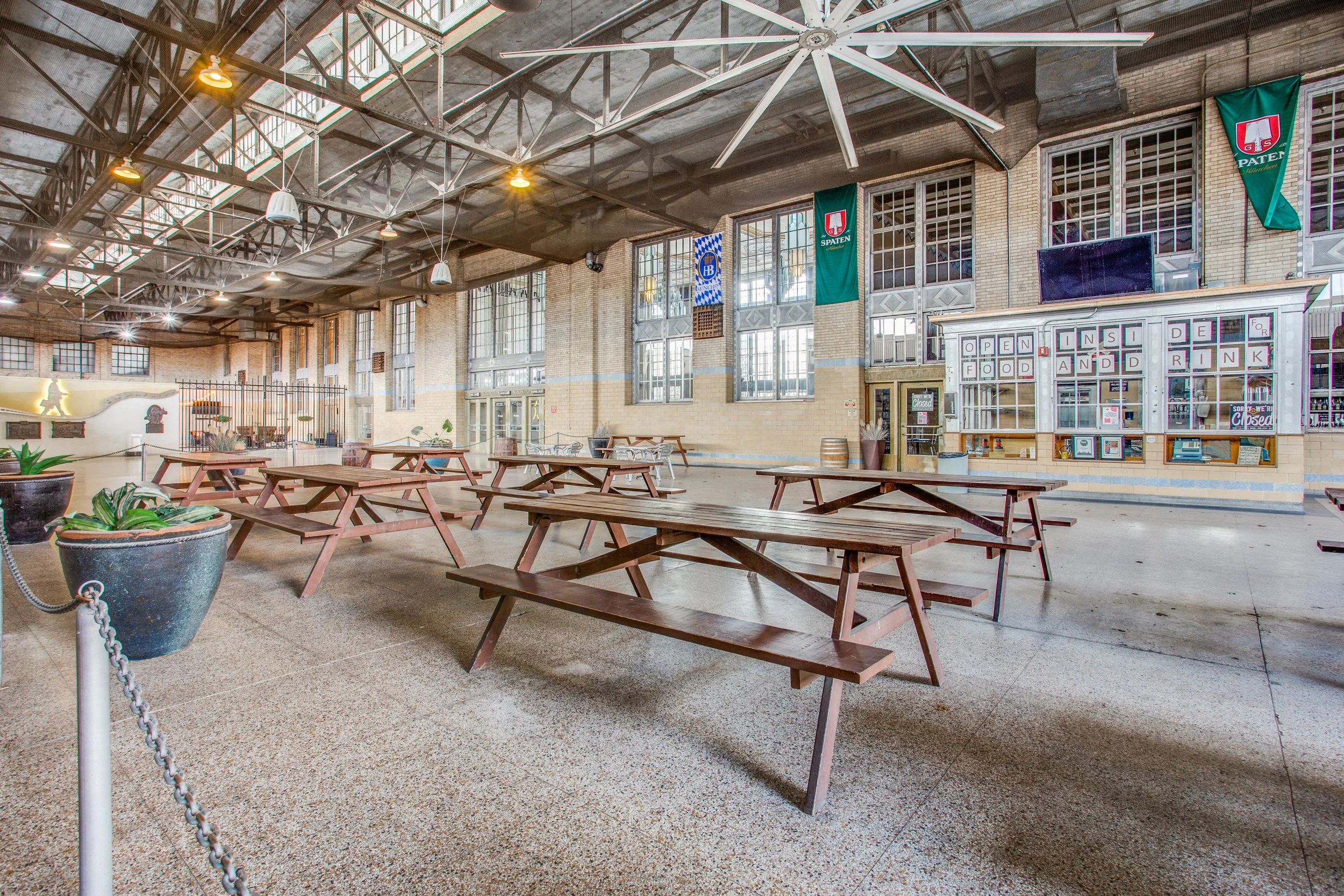 Empty indoor space with wooden picnic tables, potted plants, large windows, and banners hanging from the ceiling.