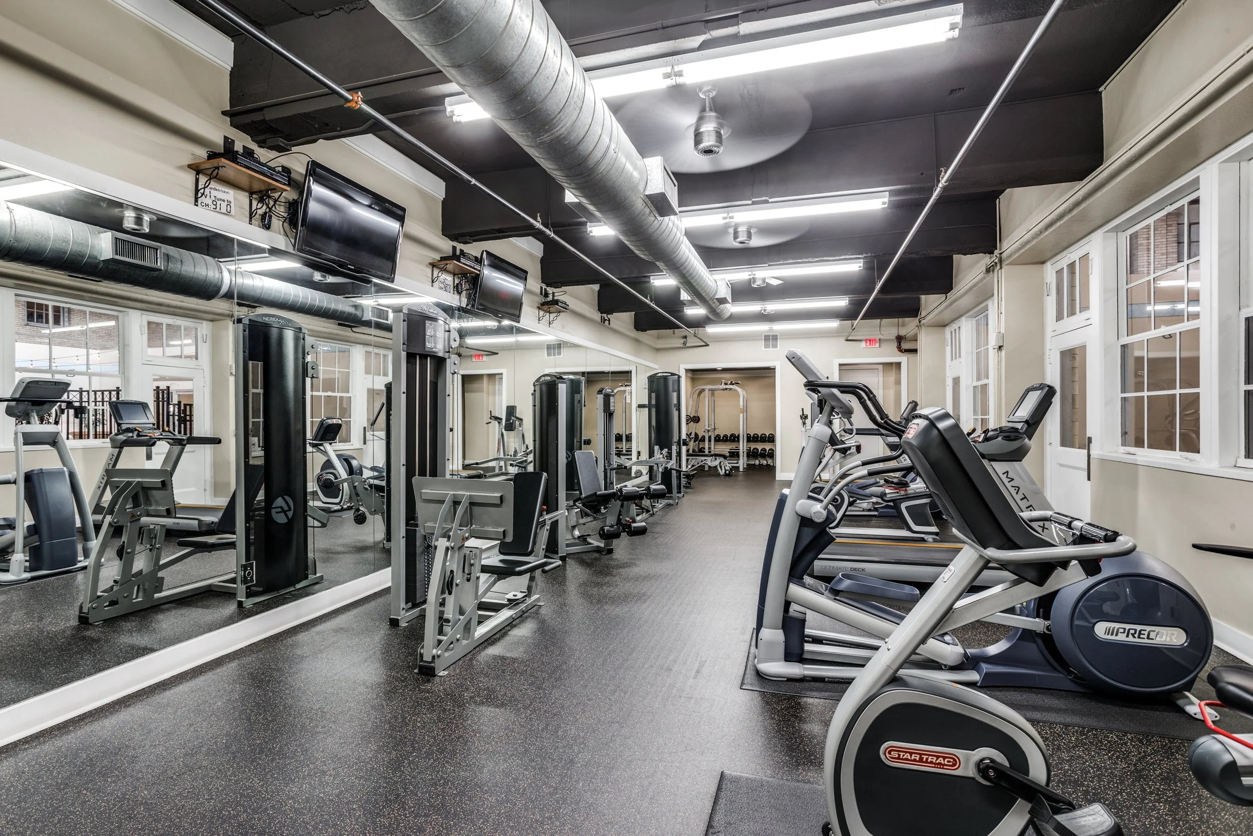 Empty gym with cardio machines in the foreground, weight stations and mirrors along the wall, ceiling with exposed ductwork, multiple ceiling lights, and windows on the right side.