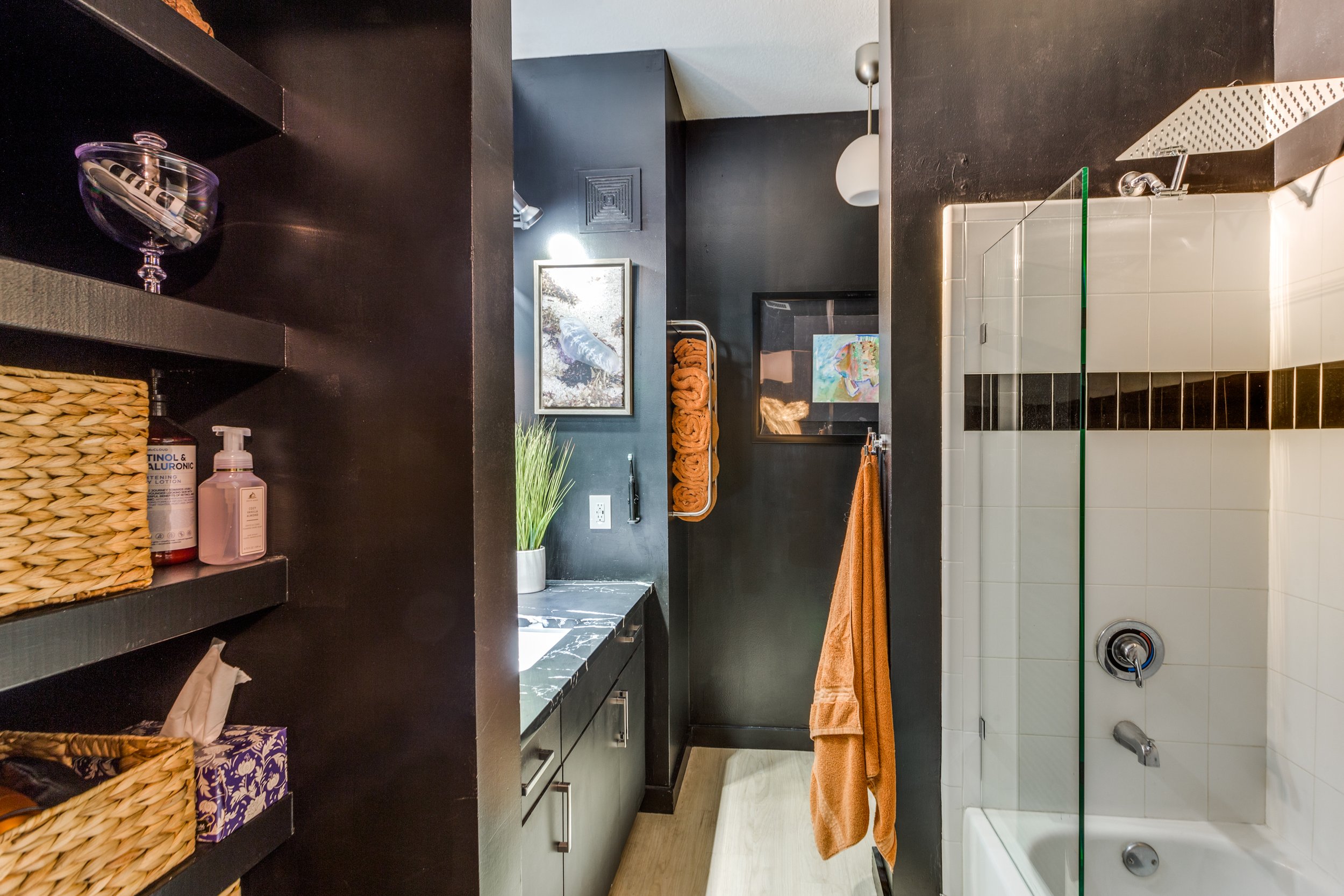 A modern bathroom with dark walls, a shower with glass door, a black and white marble countertop, and decorative towels and artwork.