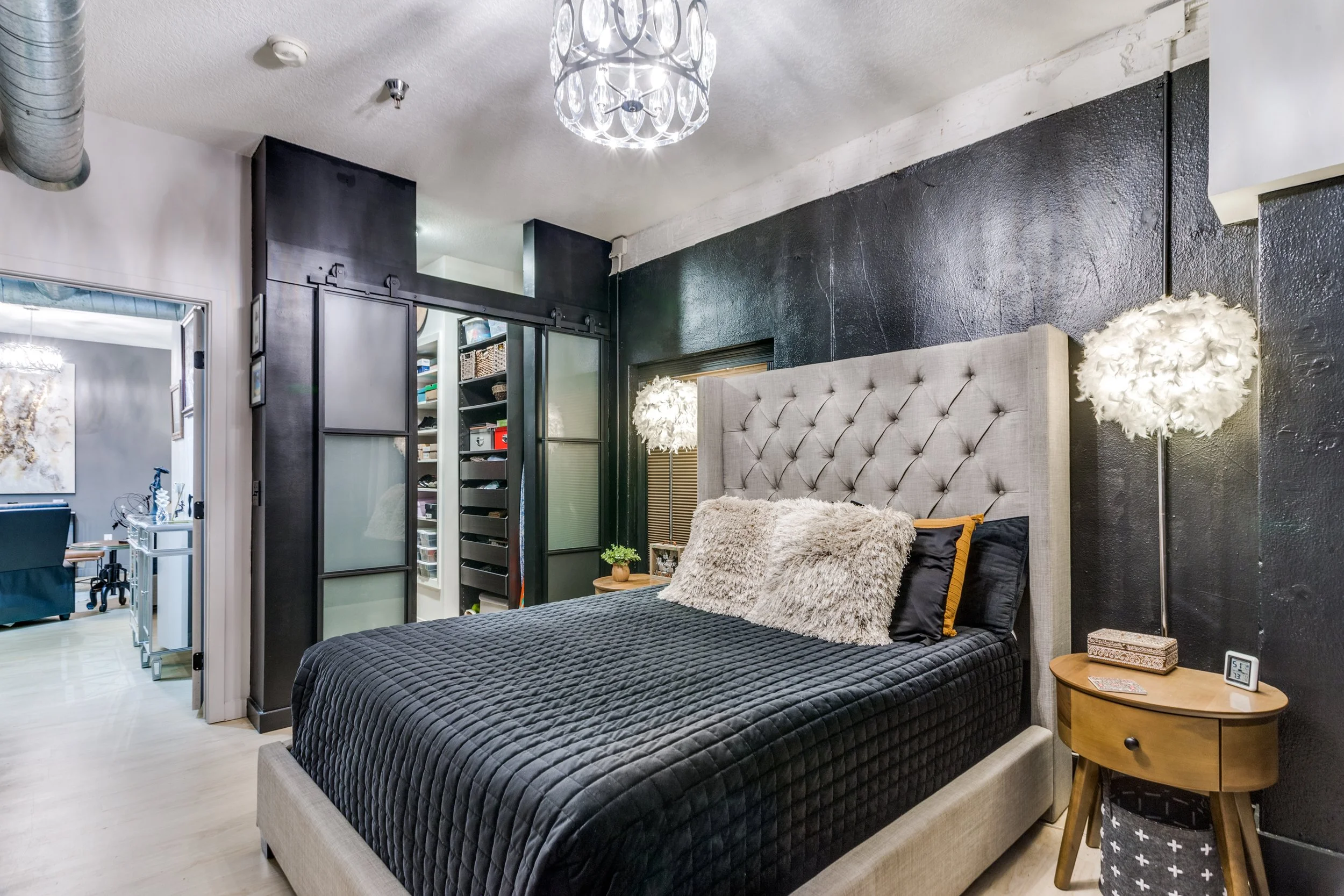 Modern bedroom with beige upholstered bed, black quilted bedding, fluffy pillows, nightstand with a small plant, lamps with furry shades, black accent wall, and adjoining walk-in closet.