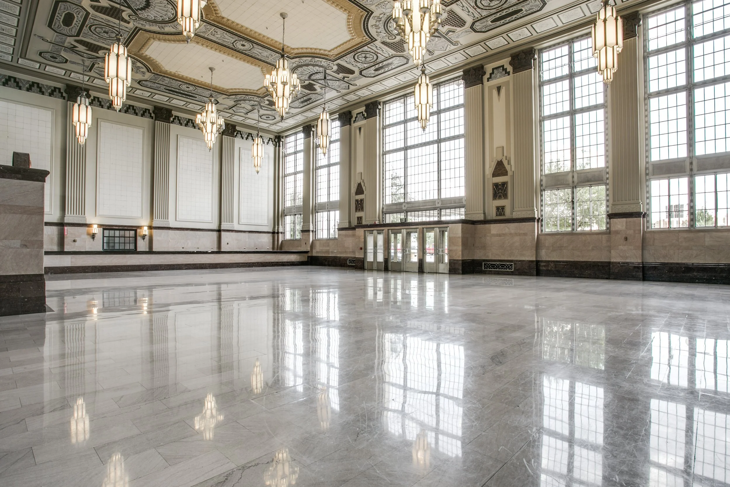 Empty elegant large hall with polished marble floors, high ceilings, large windows, and hanging chandeliers.