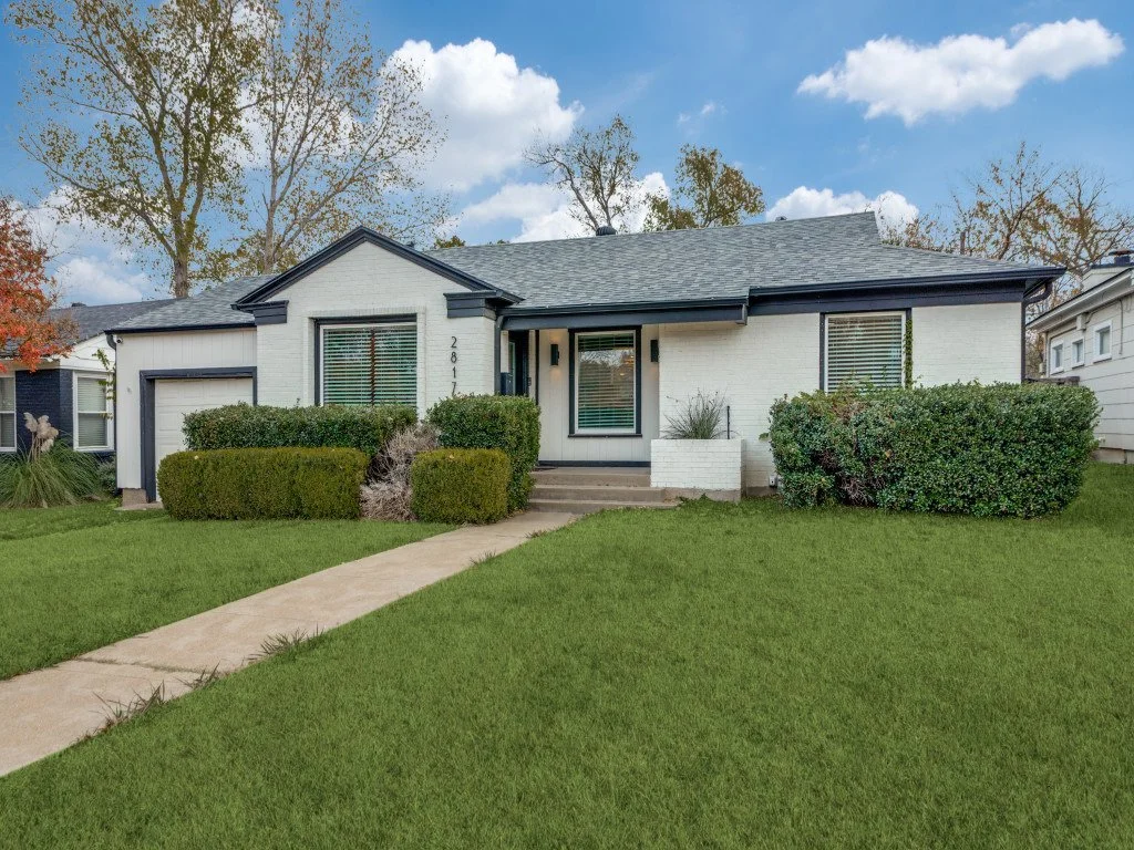 A white single-story house with black trim, a gray roof, and a landscaped front yard with a sidewalk, bushes, and green grass, under a partly cloudy sky.