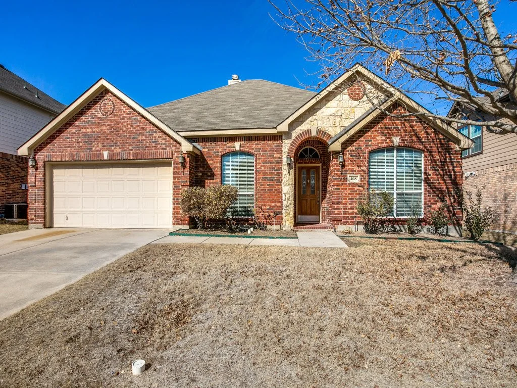 A brick house with a two-car garage, front door, and surrounding lawn and shrubbery under a clear blue sky.