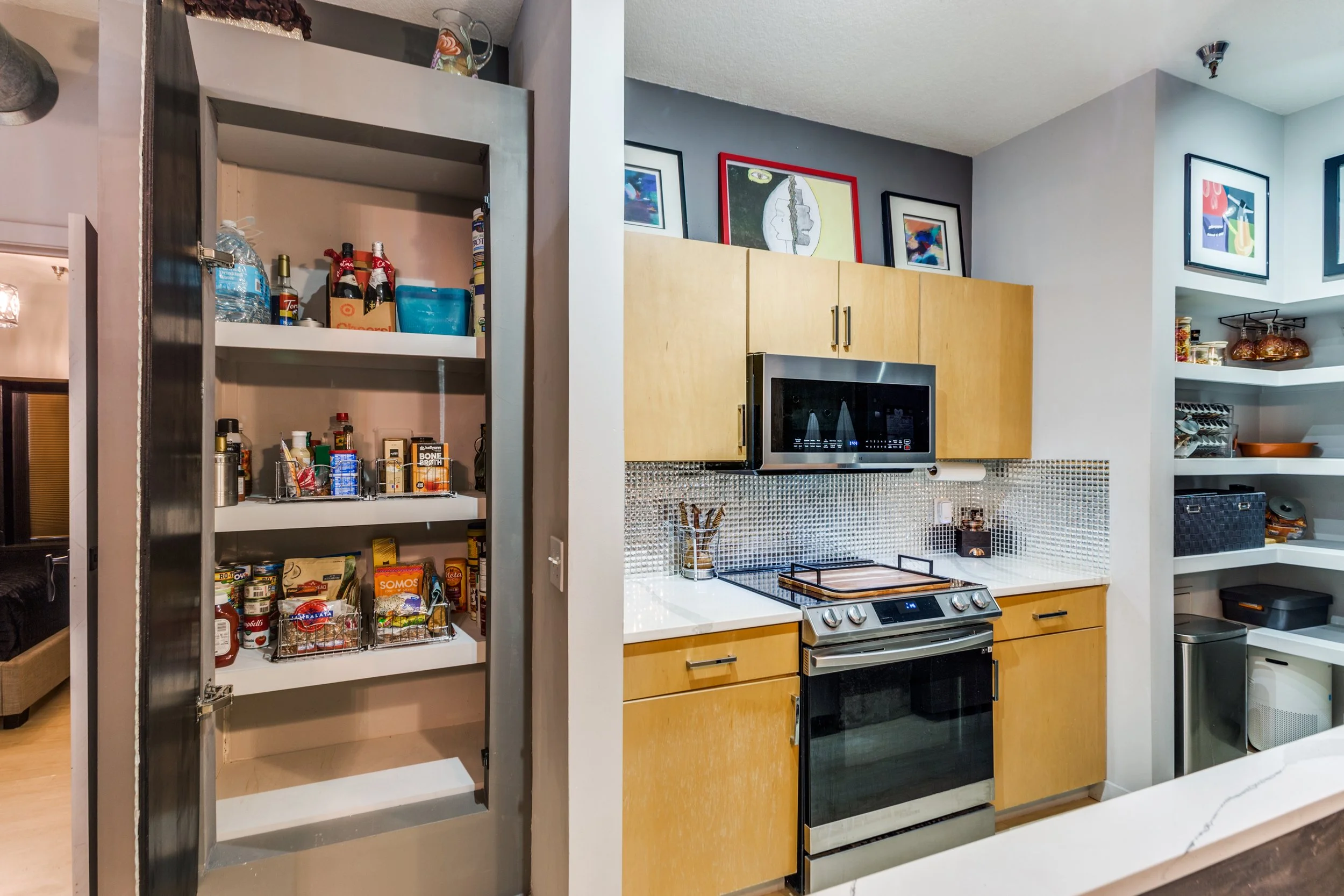 Kitchen with open pantry door on the left filled with canned foods, bottles, and snacks. In the background, there are wooden cabinets, a microwave oven, a stove, and framed pictures on the wall. On the right, there are shelves with jars, dishes, and 