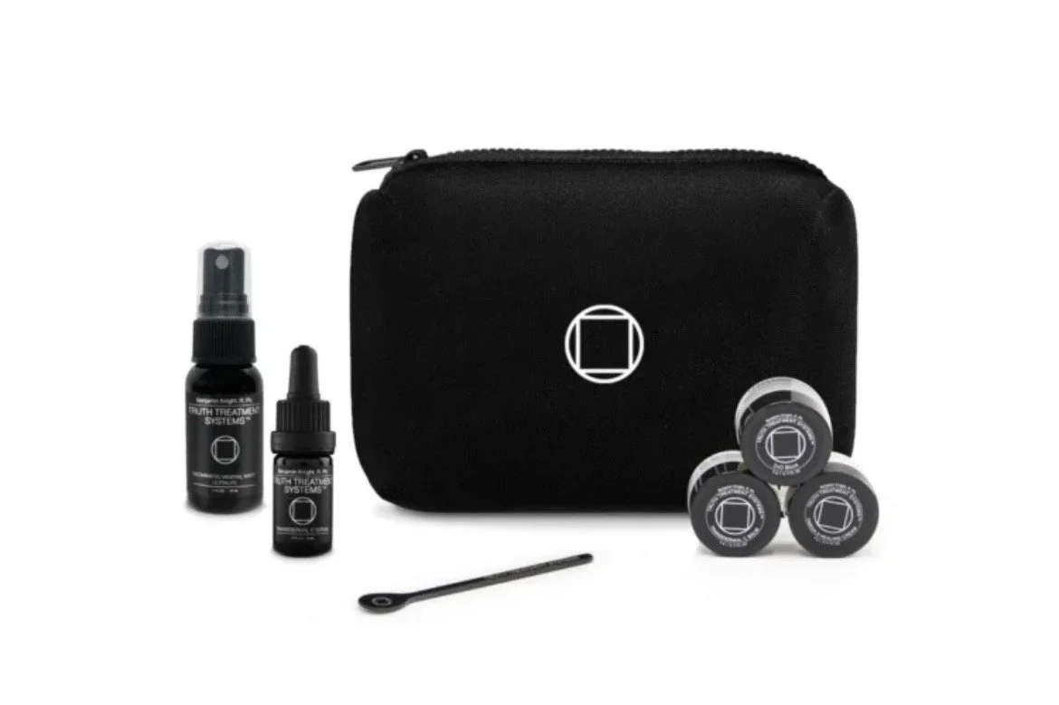 A black carrying case with a white geometric logo, surrounded by skincare products including a spray bottle, a dropper bottle, small containers, and a black applicator stick, all on a white background.