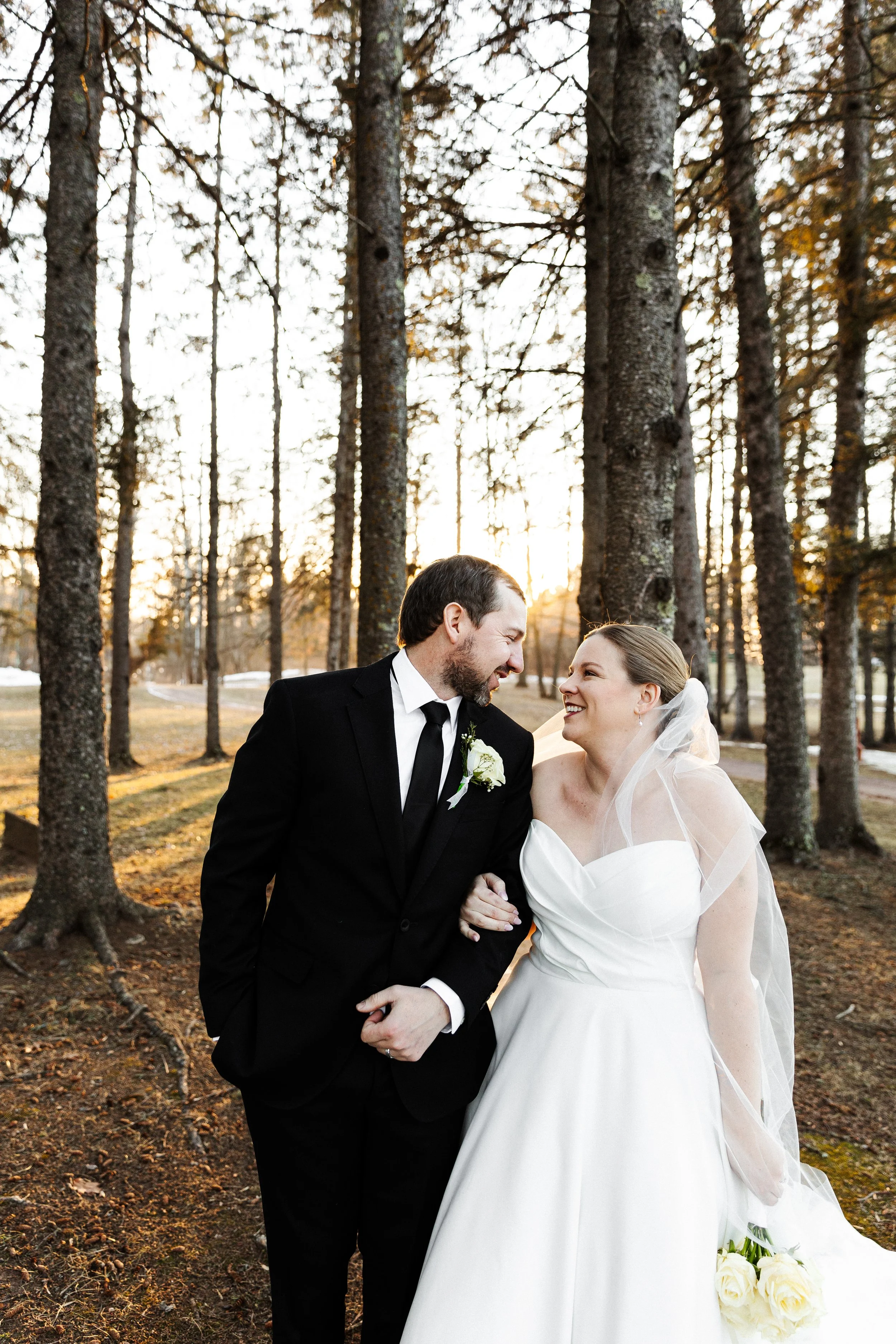 A bride and groom smile at each other in a forest during sunset, dressed in wedding attire.