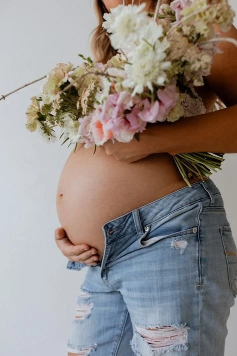 A pregnant woman holding a bouquet of white and pink flowers in front of her belly, with her other hand resting on her side, wearing distressed jeans.