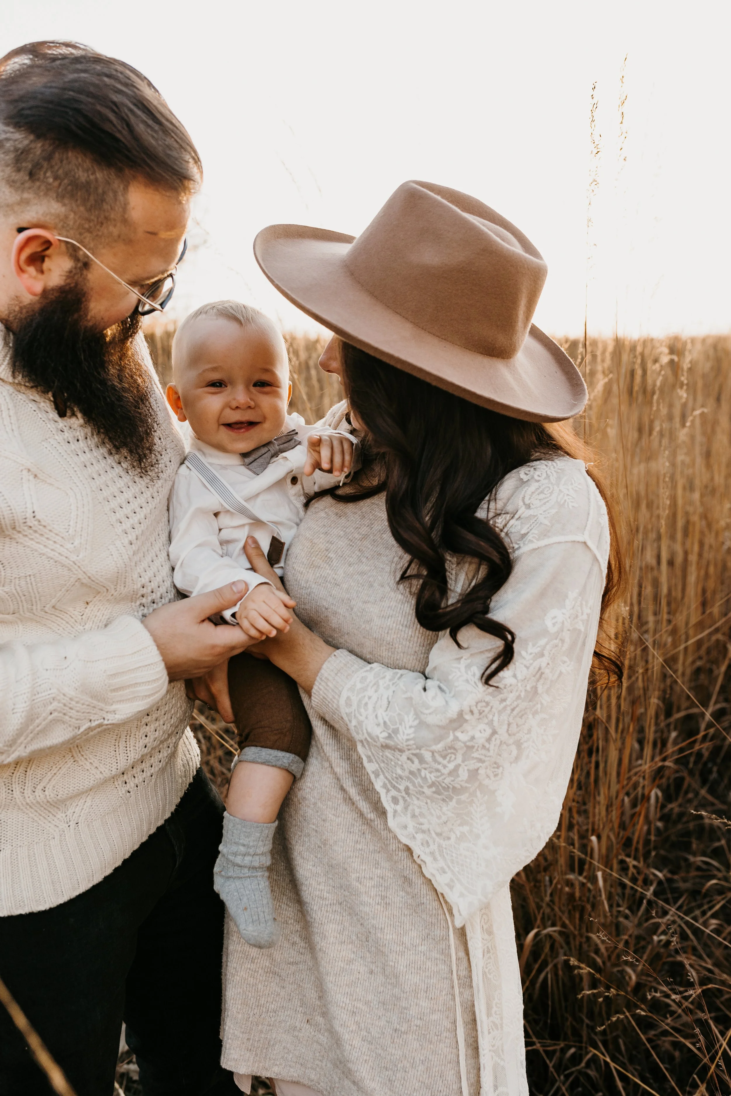 A family of three outdoors during sunset in a field of tall grass, with a bearded man, a woman wearing a hat, and a smiling baby.