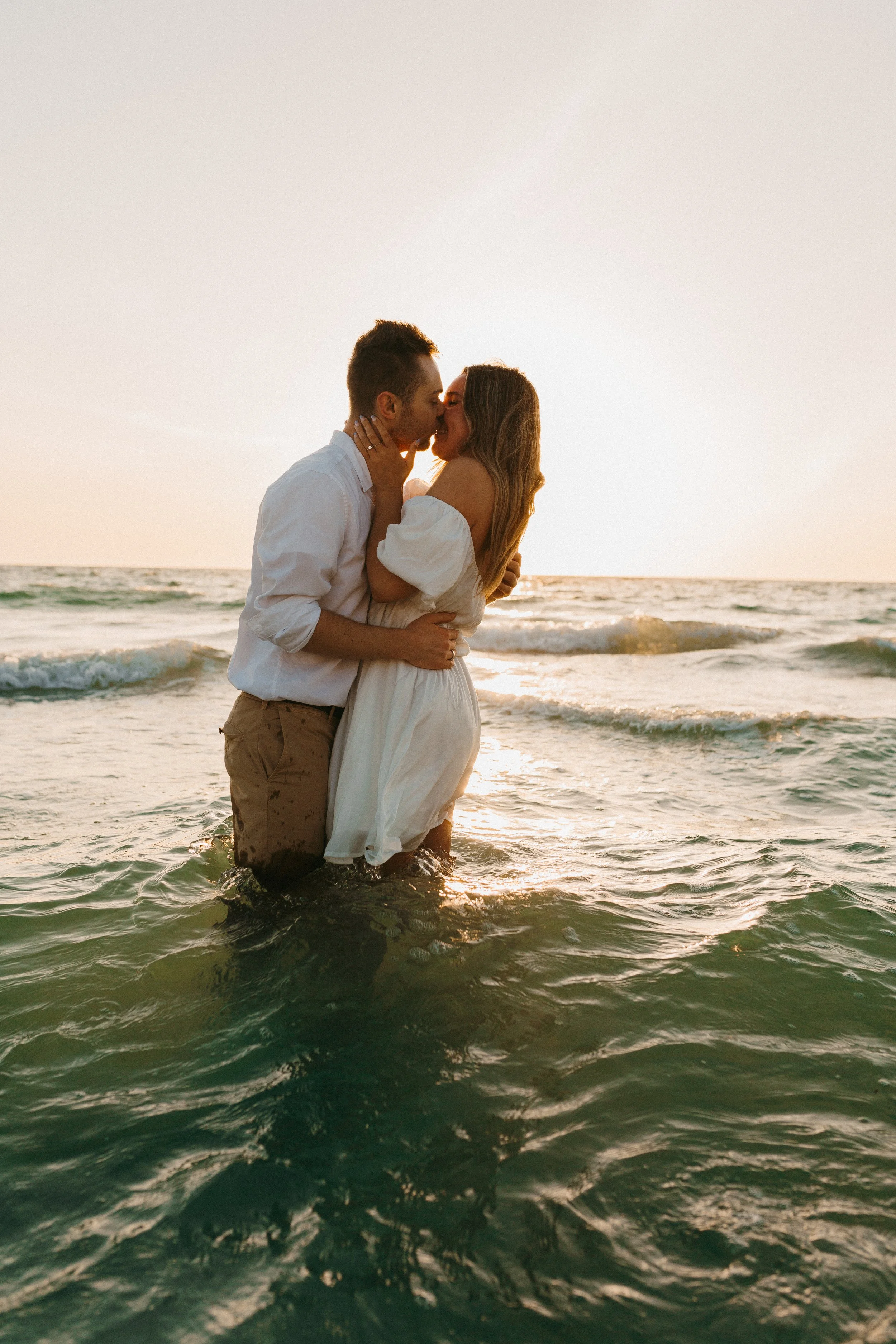 A couple wearing white clothing embracing and kissing in shallow ocean water during sunset.