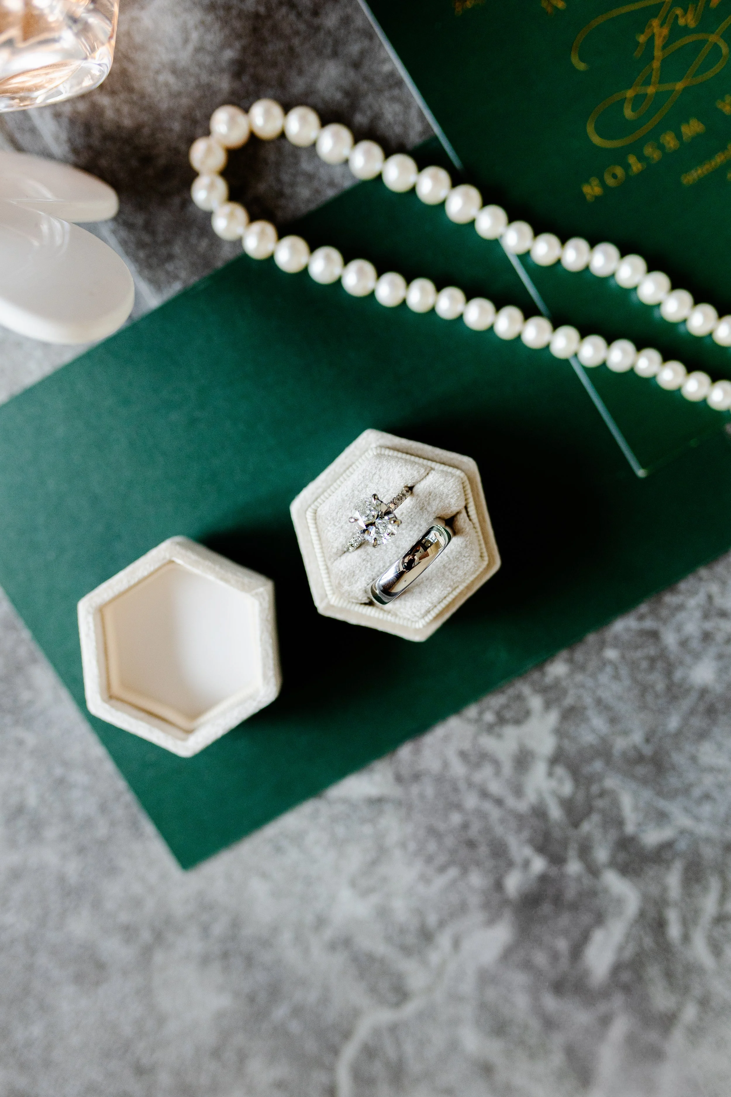 A pearl necklace, a pair of rings in a jewelry box, and a flower-shaped diamond ring are arranged on a green surface, with a gray textured background.