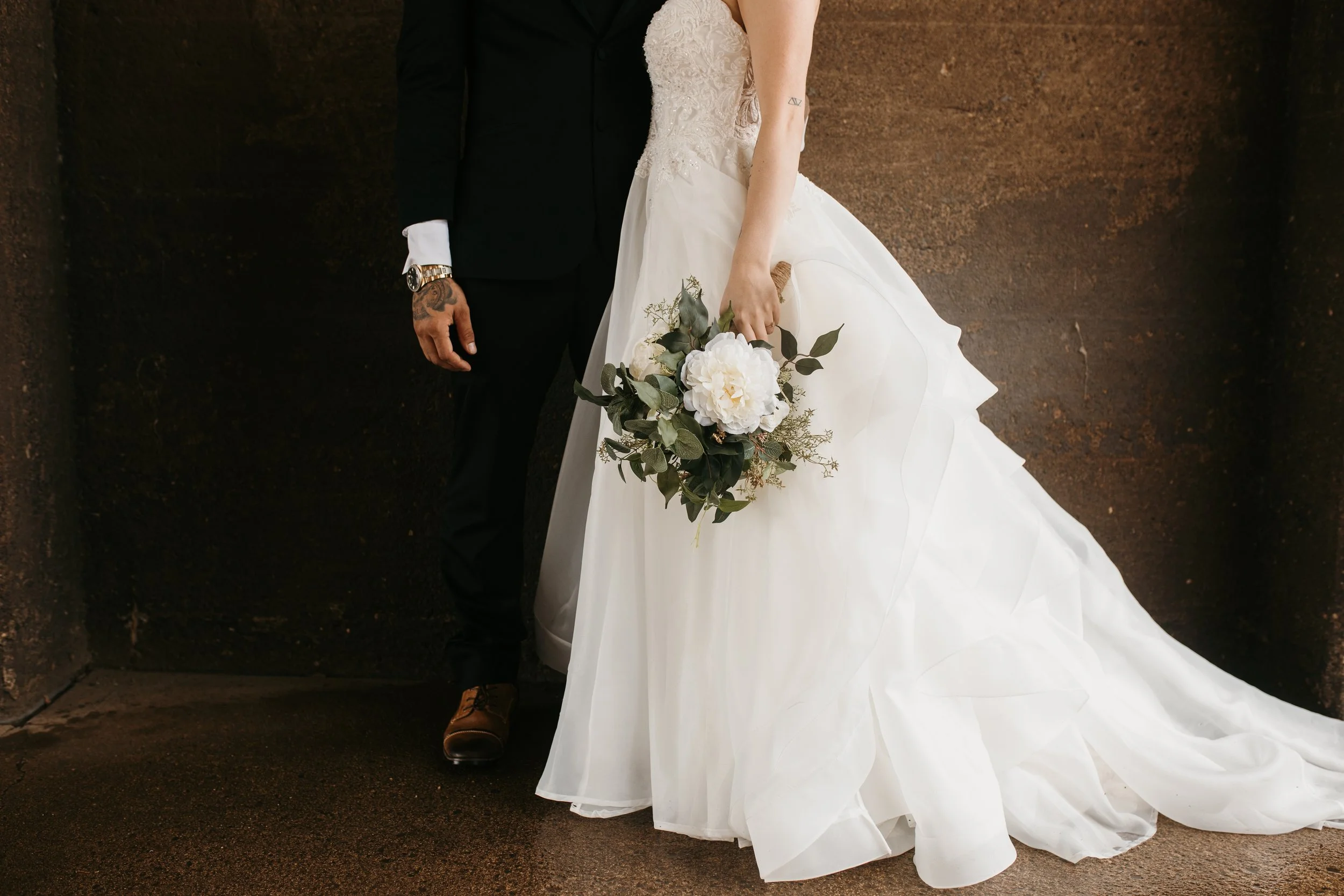 Close-up of a bride and groom from waist down, standing against a dark, textured background. The bride is wearing a white wedding gown and holding a bouquet, while the groom is dressed in a black suit with brown shoes, a watch, and a tattoo on his ha