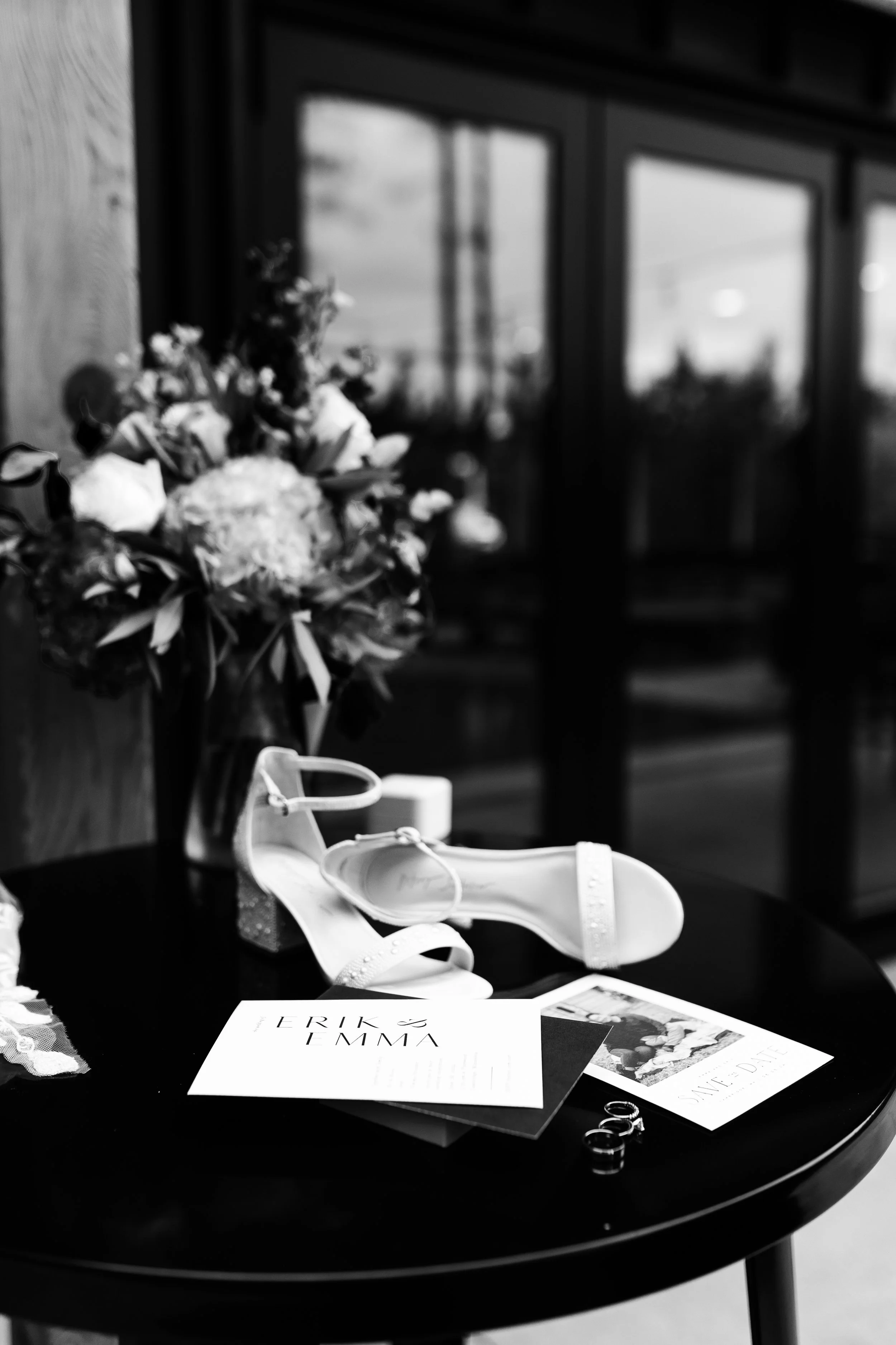 A table with a bouquet of flowers, a pair of white high-heeled shoes, a white candle, wedding rings, and wedding invitations on a black round table. In the background, there is a window with reflections.