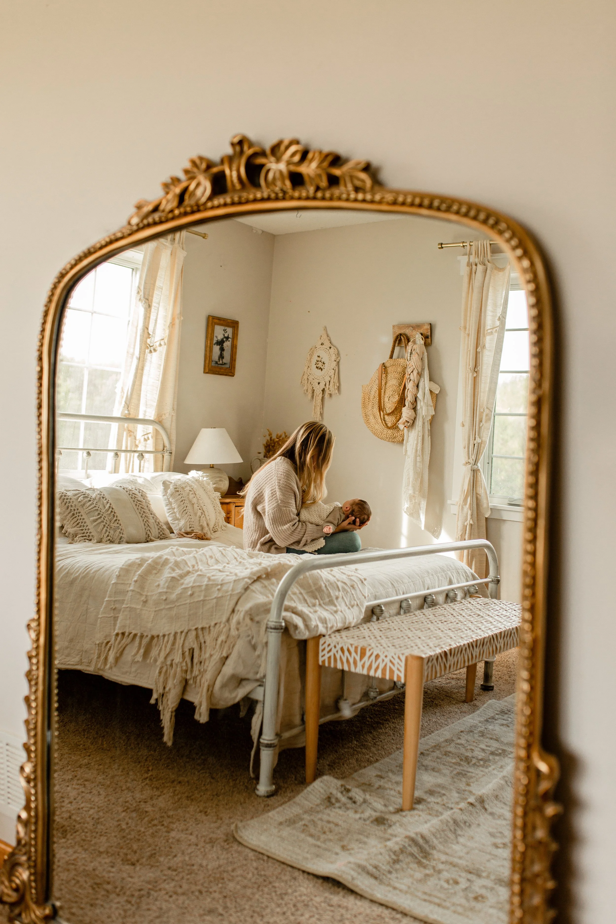 A woman holding a newborn baby is reflected in an ornate gold-framed mirror, with a cozy bedroom scene including a bed, window, and wall decor visible in the background.