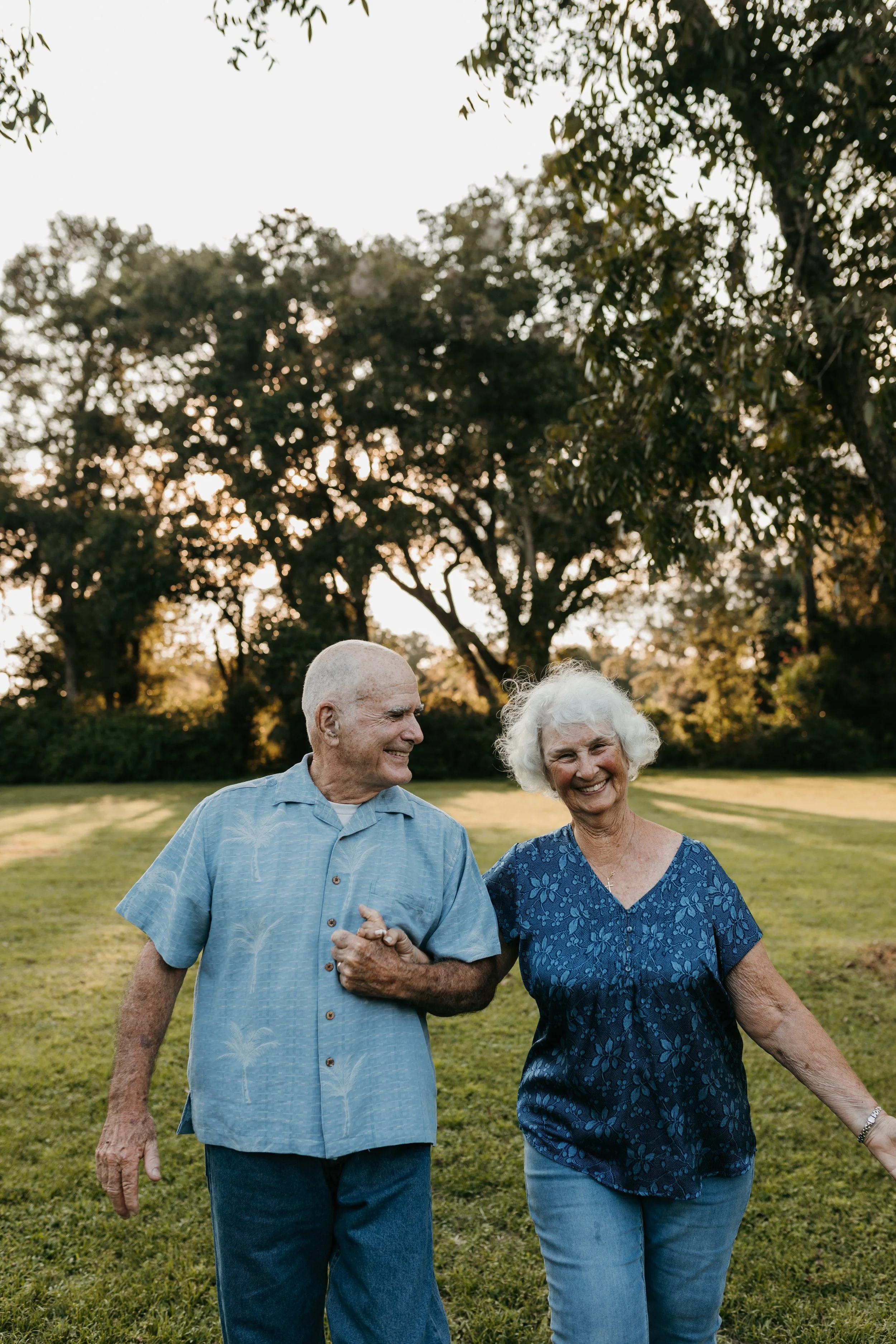 An elderly couple walking together outdoors on a grassy field, smiling and holding hands, with tall trees and sunlight in the background.