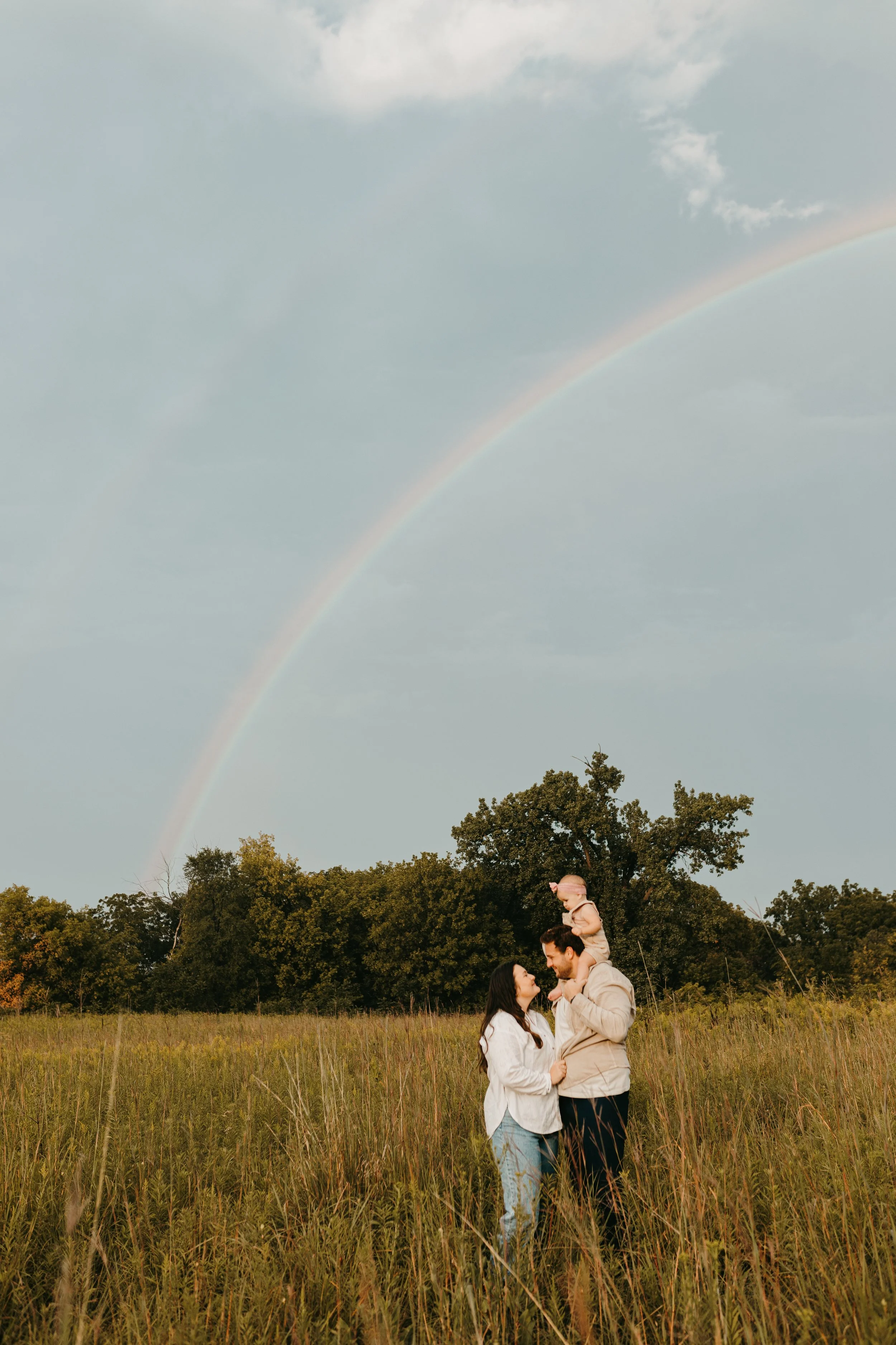 A family of three standing in a grassy field under a rainbow, with trees in the background and a cloudy sky.