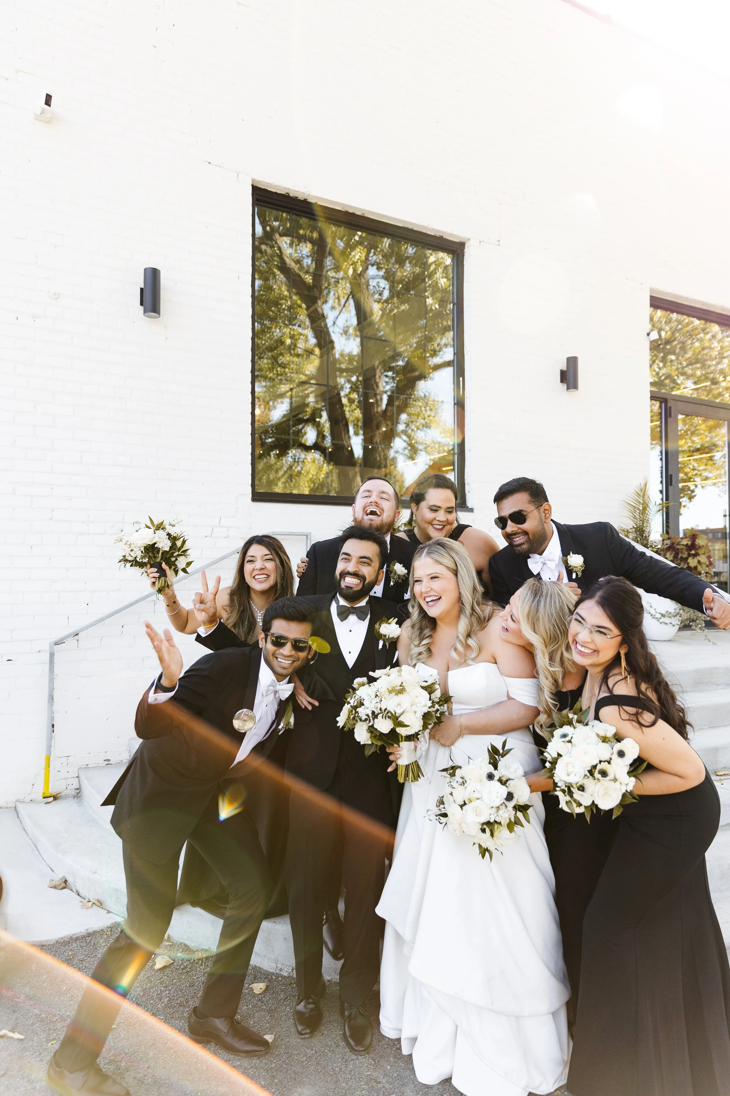 A diverse group of wedding guests and bride and groom smiling and celebrating outside a modern white brick building, with some making peace signs and holding bouquets of white flowers.