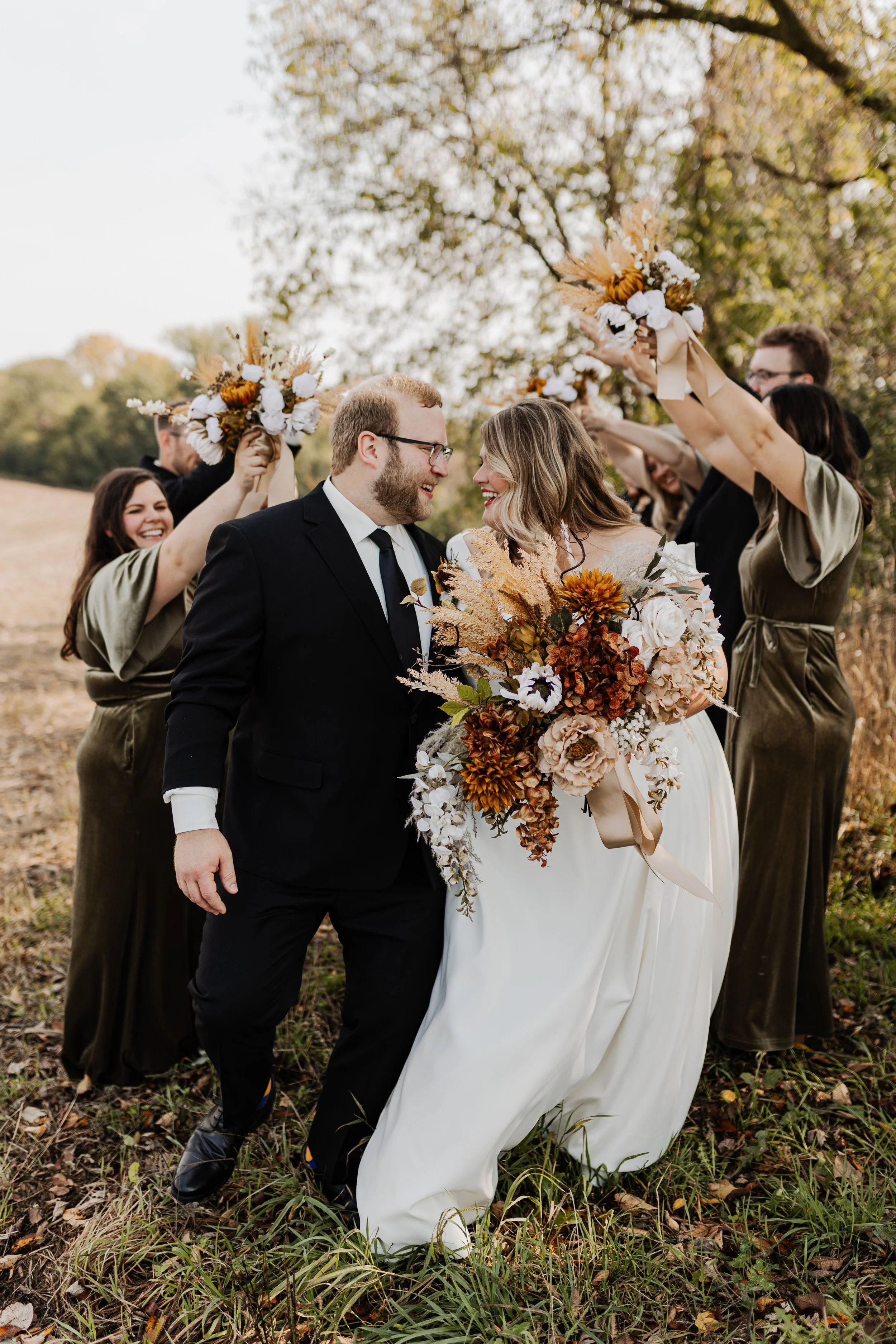 A newlywed couple celebrating outdoors with friends holding bouquets of flowers. The bride wears a white gown and holds a large bouquet; the groom wears a black suit. Friends raise bouquets in a festive toast against a backdrop of trees with autumn l