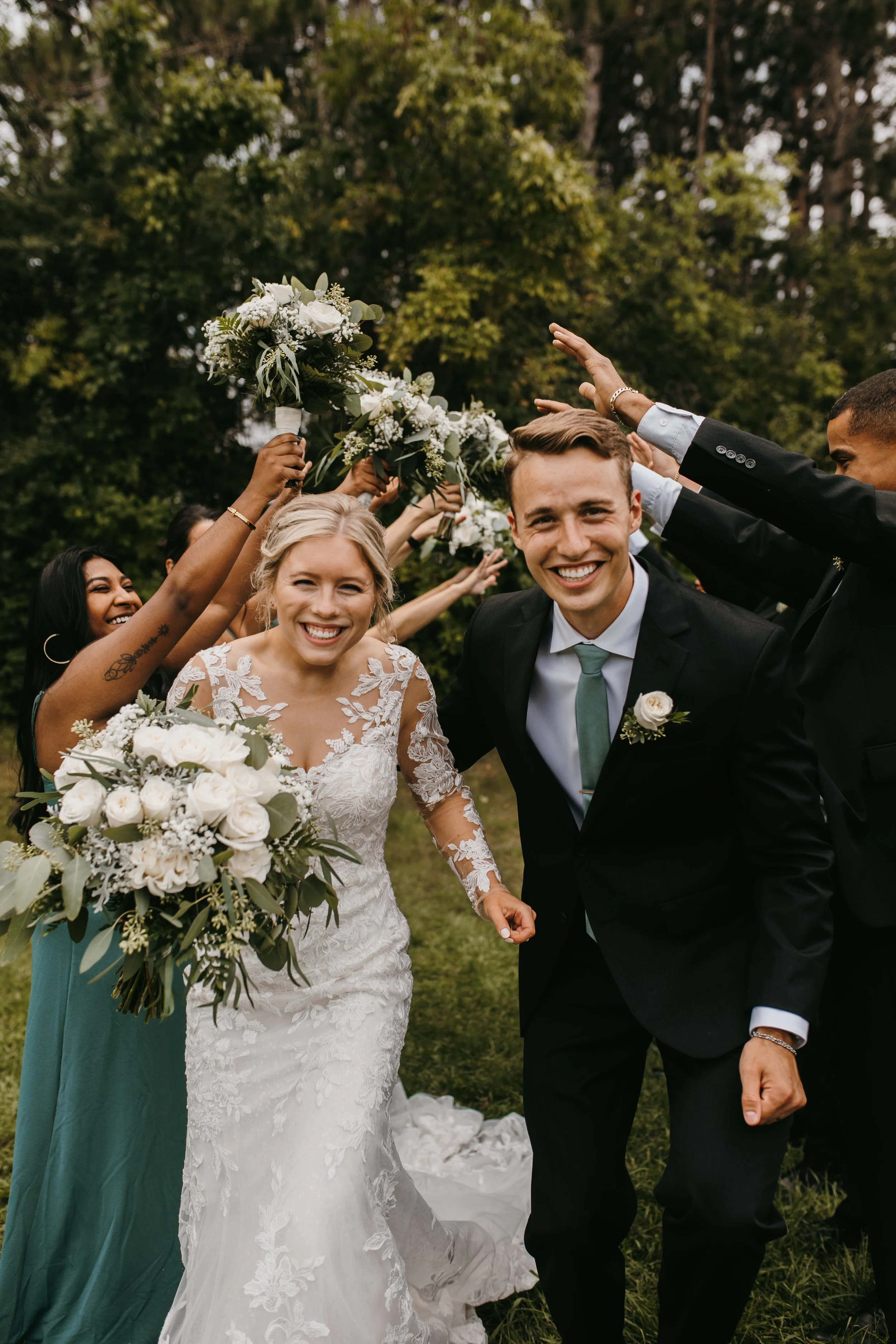 A newly married couple in wedding attire celebrating with friends outdoors, smiling happily, with friends holding bouquets and making a celebratory arch behind them.