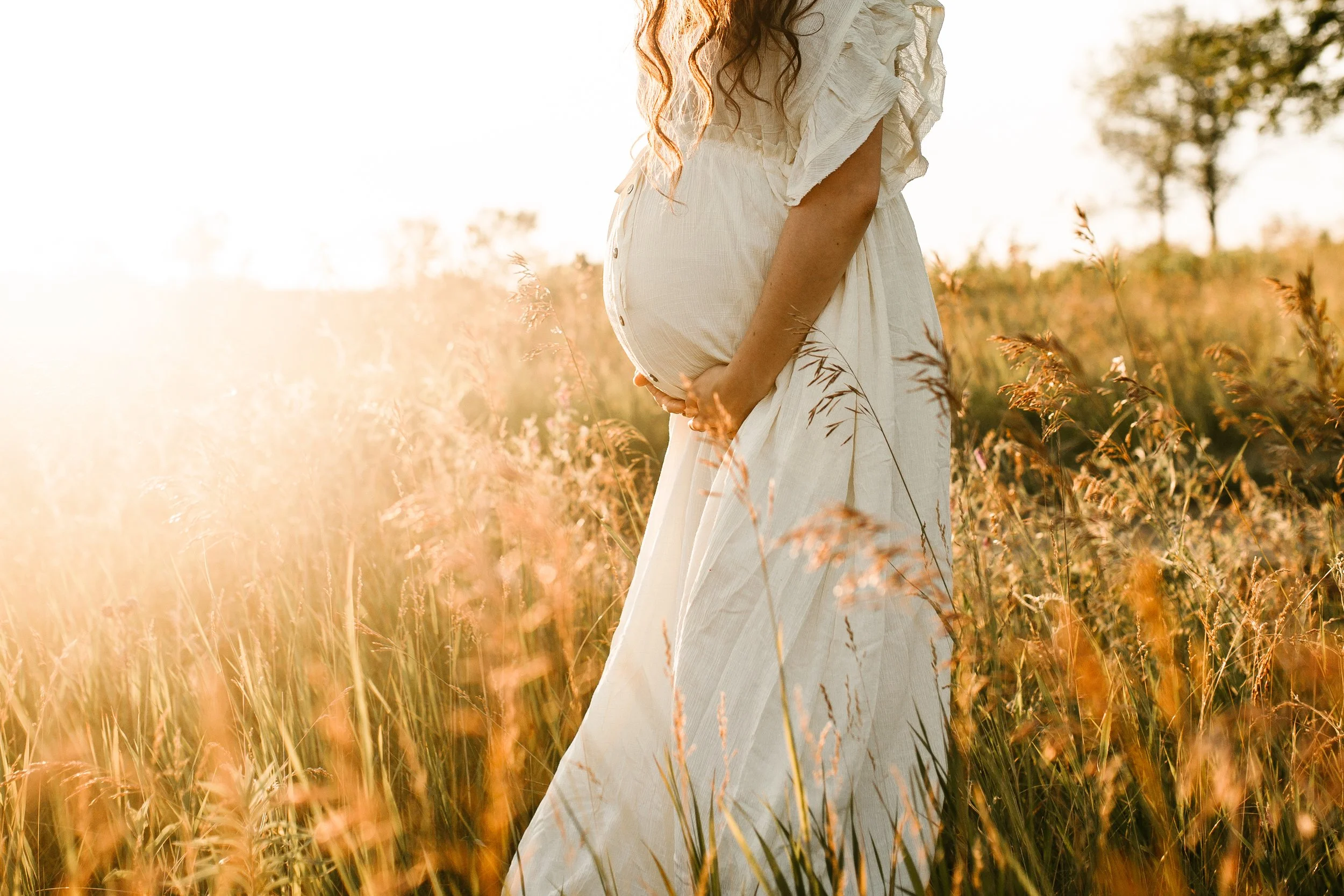 Pregnant woman in a flowing white dress standing in a field of tall grass during sunset.