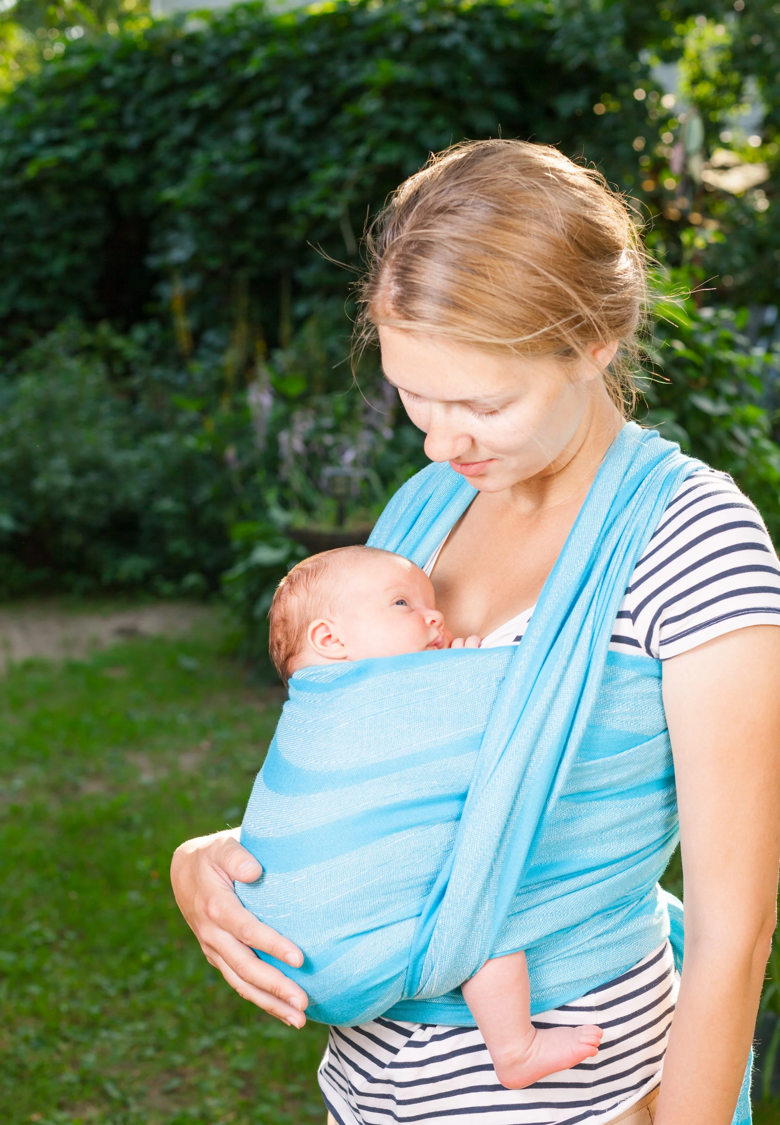 White woman with light coloured hair looking down lovingly at her baby, who is being worn in a woven baby wrap in a front wrap cross carry. They are outdoors with greenery in the background.