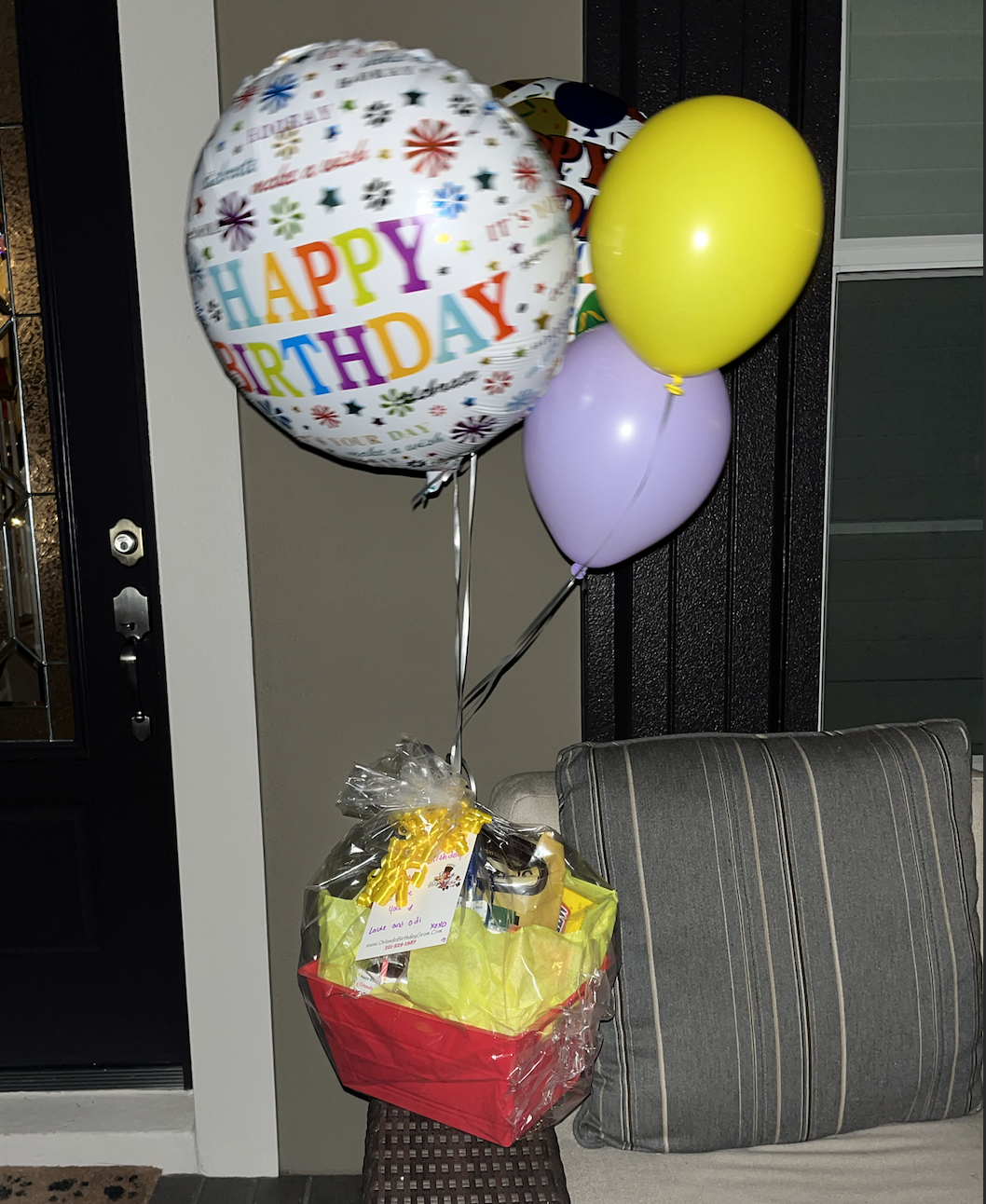 Cluster of colorful balloons, including a large 'Happy Birthday' balloon, hanging above a gift basket wrapped in cellophane, with a striped cushion on a brown seat nearby.