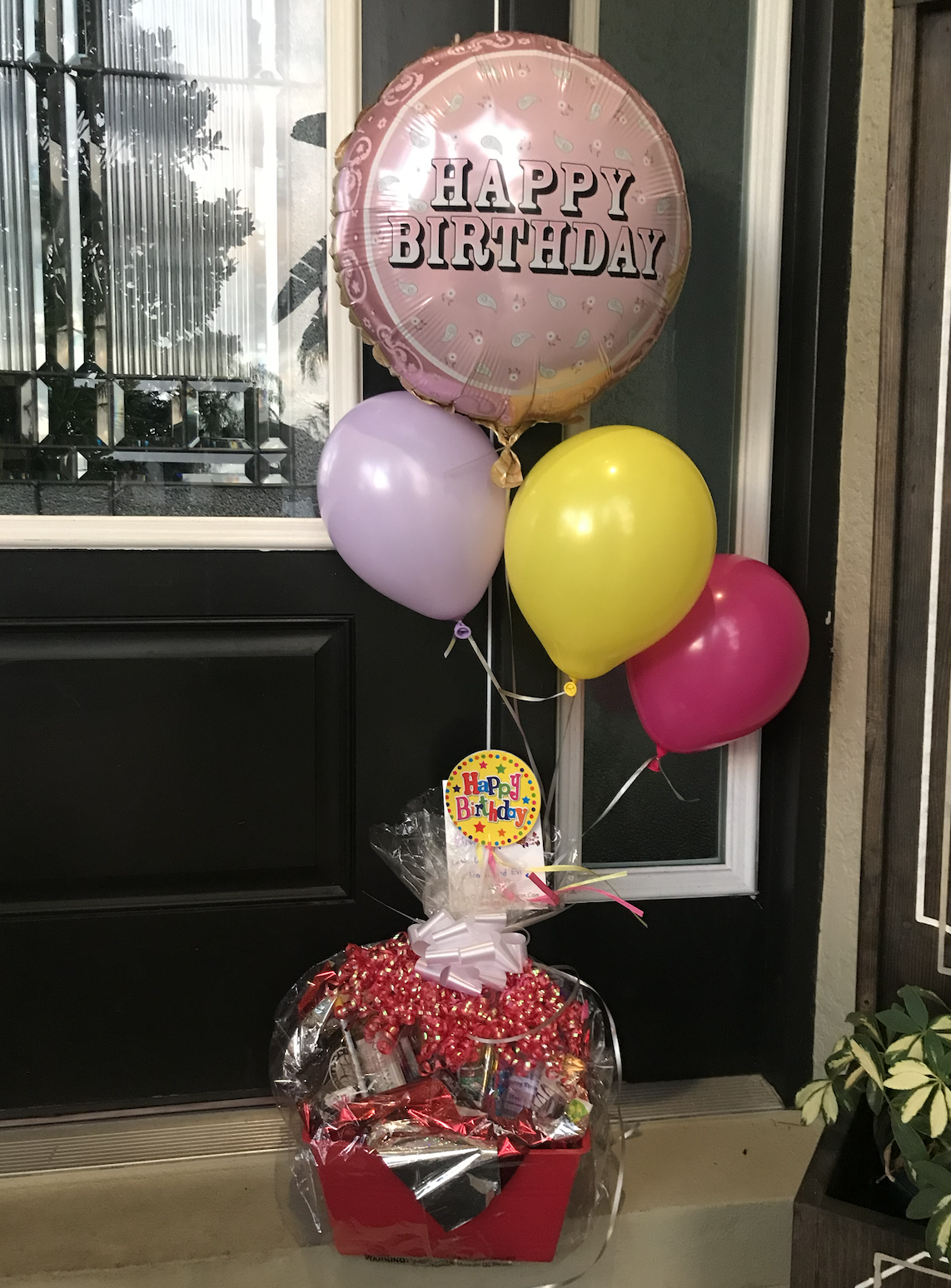 Birthday decoration with a balloon bouquet featuring a large pink 'Happy Birthday' balloon, two smaller purple and yellow balloons, and one pink balloon, all attached in a gift basket wrapped in clear plastic with red accents and a colorful 'Happy Bi