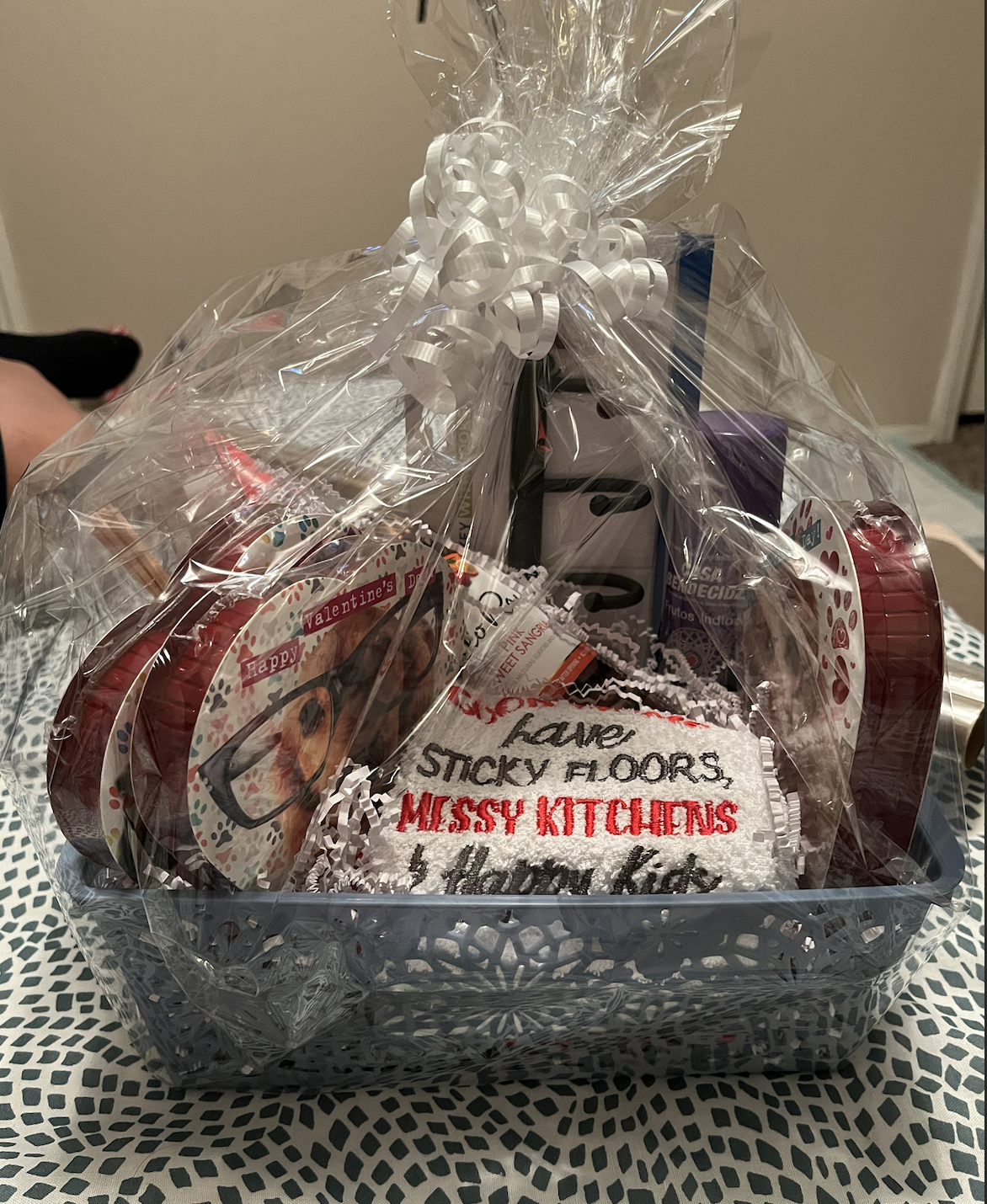 Gift basket wrapped in clear plastic with decorative bow, containing Valentine's themed items and a cleaning cloth with text that reads, 'Have sticky floors, messy kitchens &...