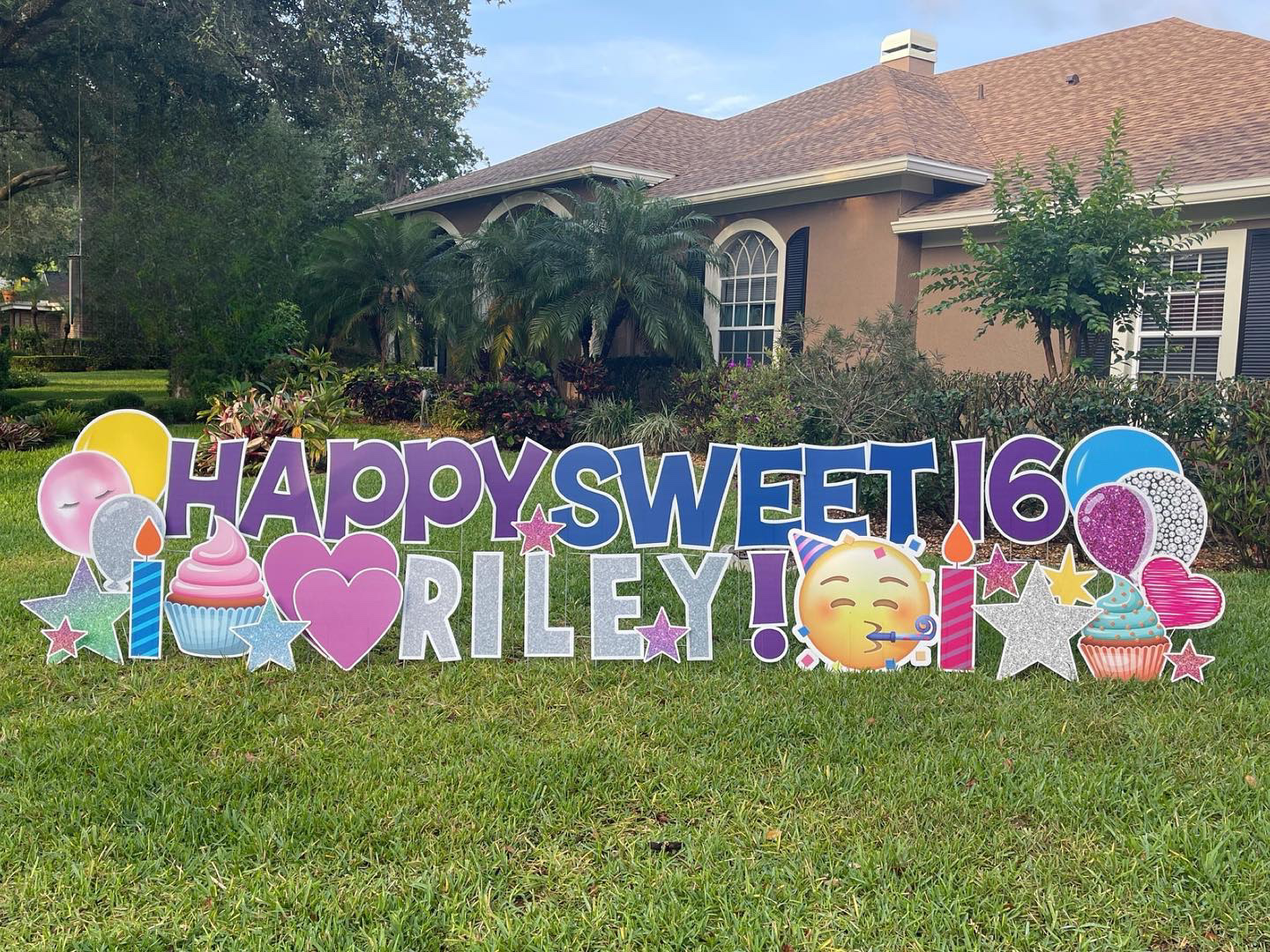 Colorful birthday yard sign with balloons, cupcakes, and emoji, celebrating Riley's 16th birthday in front of a house with a garden.