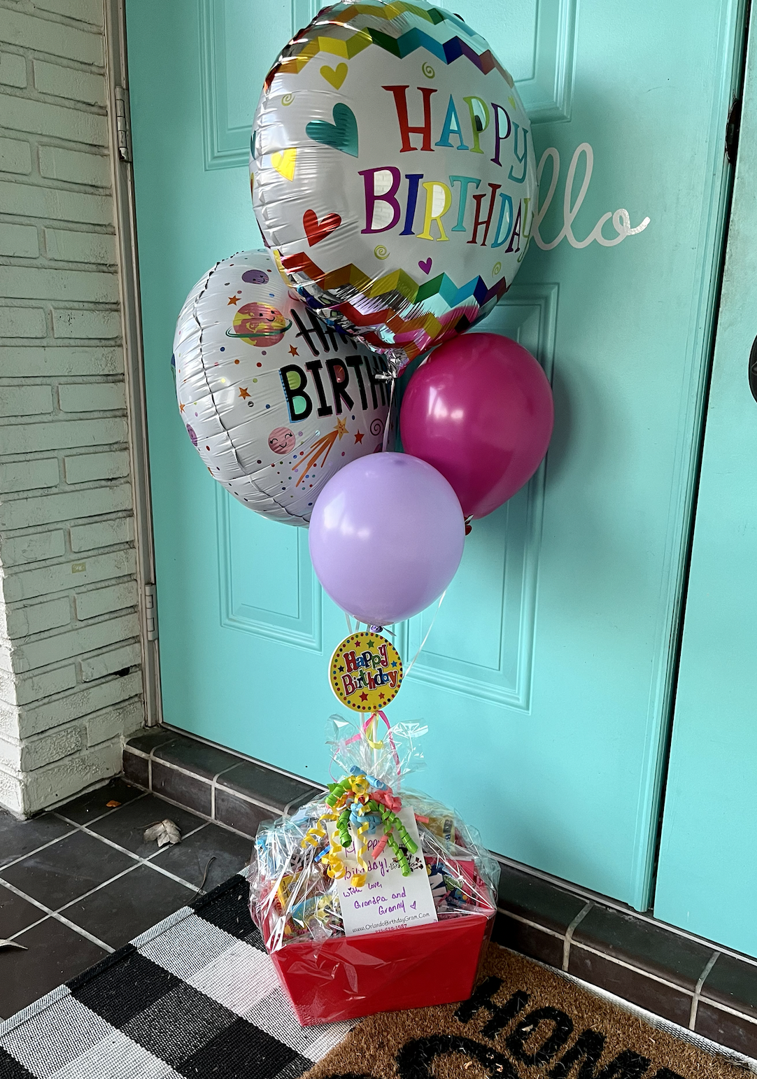 A colorful birthday gift arrangement with balloons, including two large round balloons with birthday messages, one pink, one lavender, and a smaller round balloon with a 'Happy Birthday' sticker. The balloons are anchored in a gift basket wrapped in 