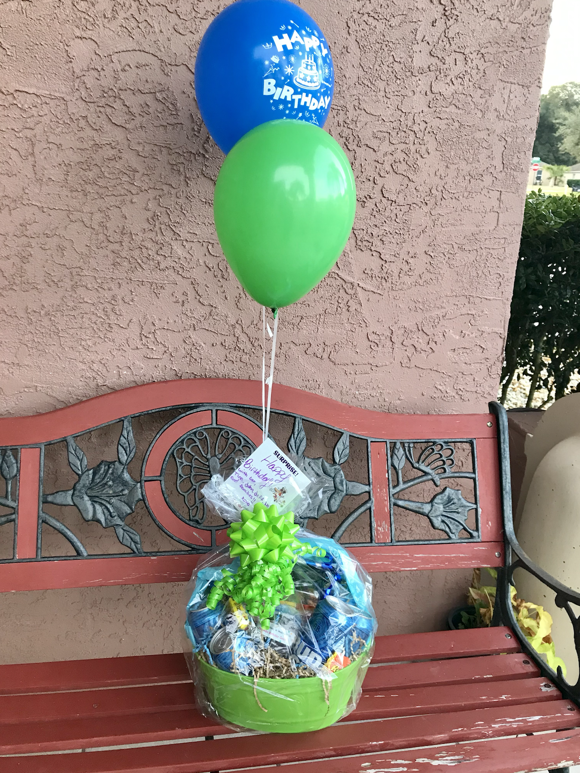 Birthday gift basket wrapped in cellophane with a large green bow, featuring balloons—one blue with 'Happy Birthday' and a cake illustration, and one green—attached to the basket, placed on a red metal bench against a textured pink wall.