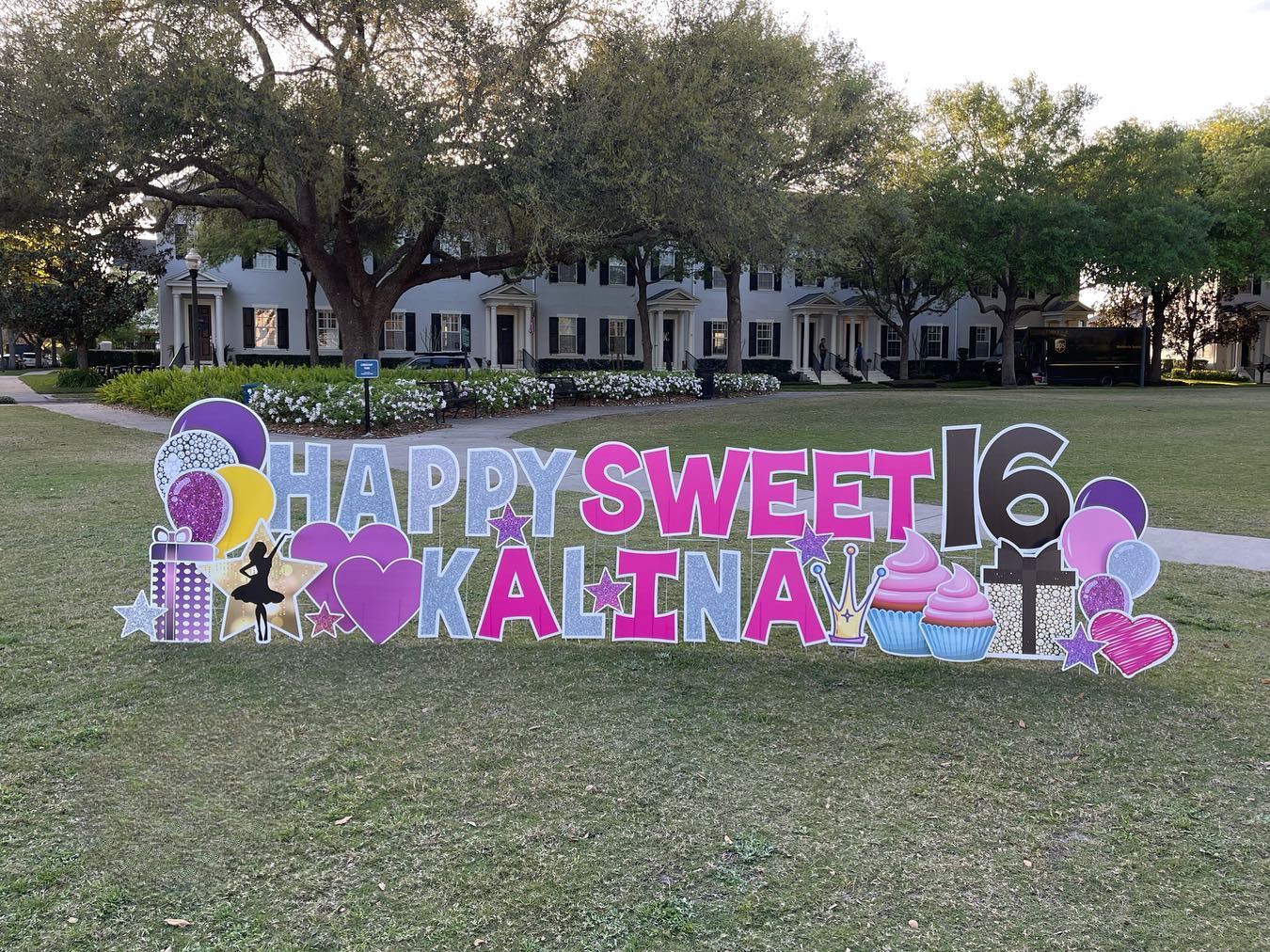 Colorful birthday sign reading "Happy Sweet 16 Kalina" with balloons, cupcakes, a crown, a ballerina, and hearts, set on a grassy area with trees and residential buildings in the background.
