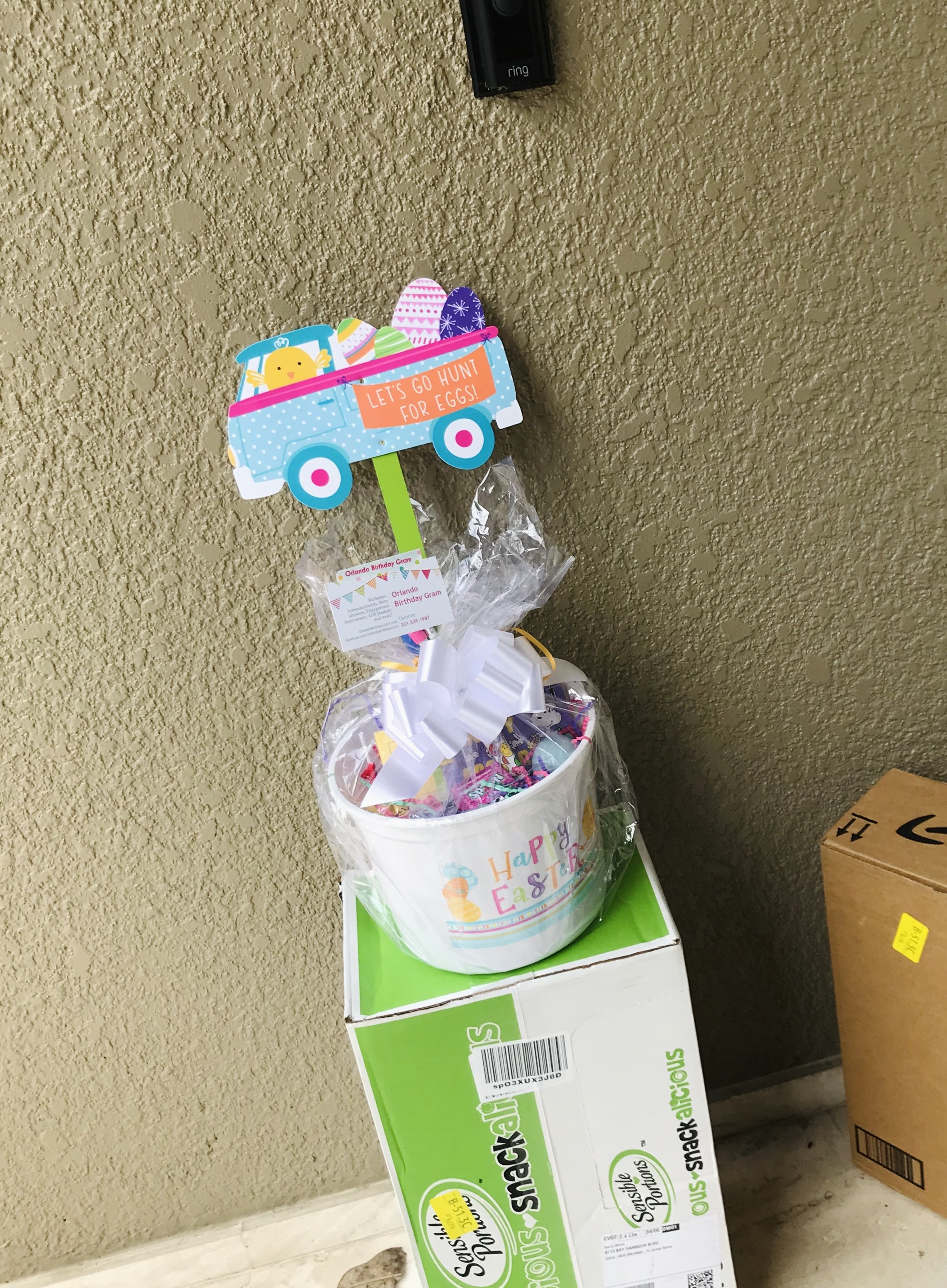 Easter themed gift basket with a sign that says 'Let's go hunt for eggs!' on top, placed on a green and white box, with a wall outlet and telephone on a beige wall in the background.