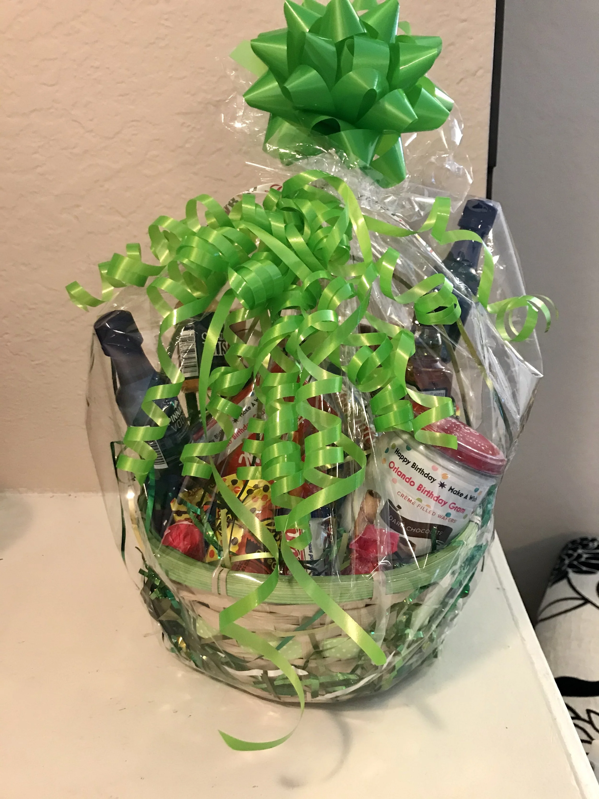 Gift basket wrapped in clear cellophane with green curling ribbons and bows, containing assorted snacks and small bottles.