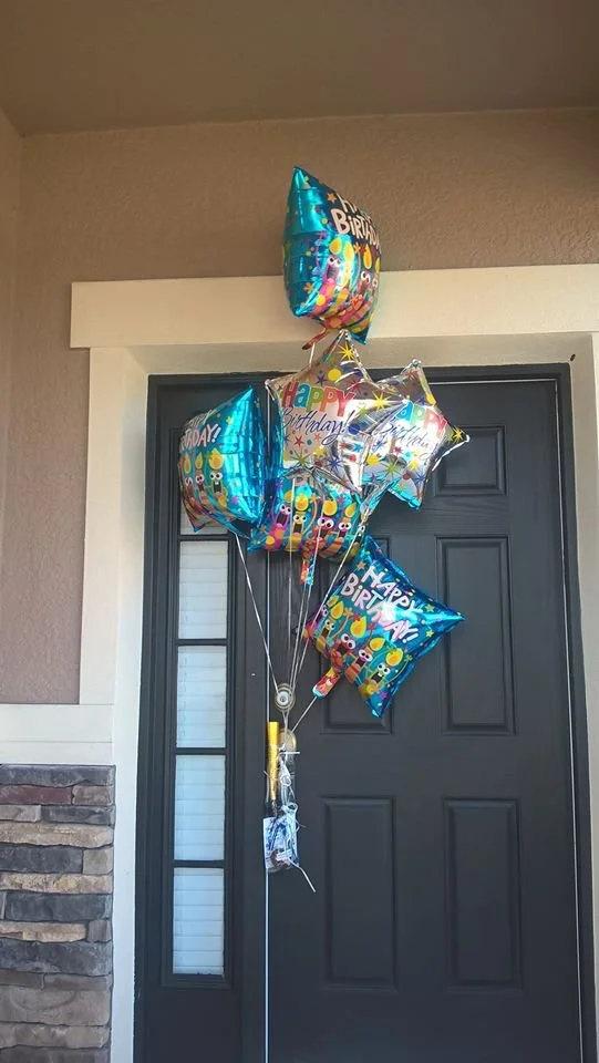 A bundle of birthday balloons, including star-shaped and rectangular helium balloons with colorful birthday-themed designs, is tied together and hanging in front of a black front door.