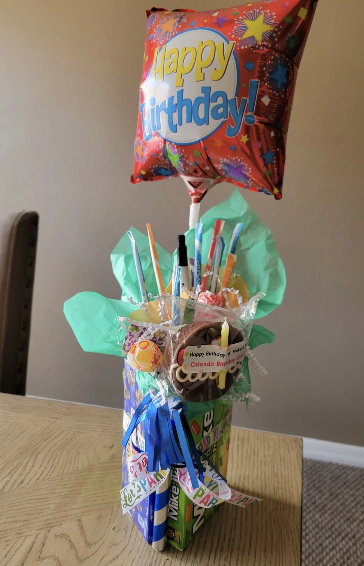 A decorated birthday gift arrangement with a 'Happy Birthday' balloon on top, colorful tissue paper, a chocolate cupcake with a candle, and various candies and straws, all in a gift bag with birthday-themed ribbons.