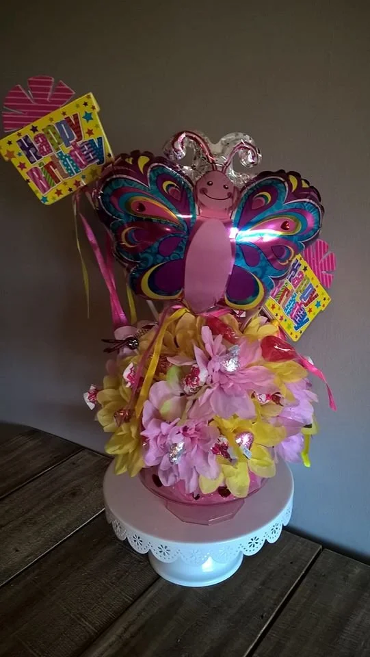 Colorful birthday gift arrangement with pink and yellow artificial flowers, a butterfly decoration with vibrant patterns, and birthday themed cards with 'Happy Birthday' message, placed on a white decorative stand.