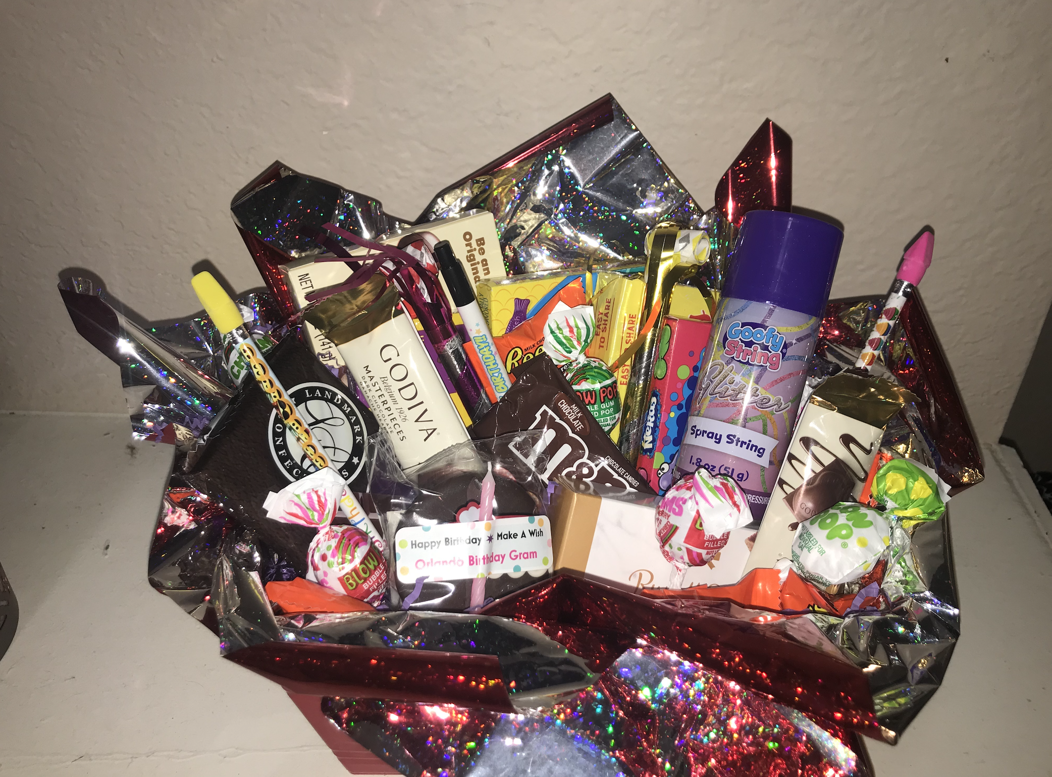 A gift basket wrapped in shiny red and silver wrapping paper, filled with various candies, snacks, and a toy spray string.