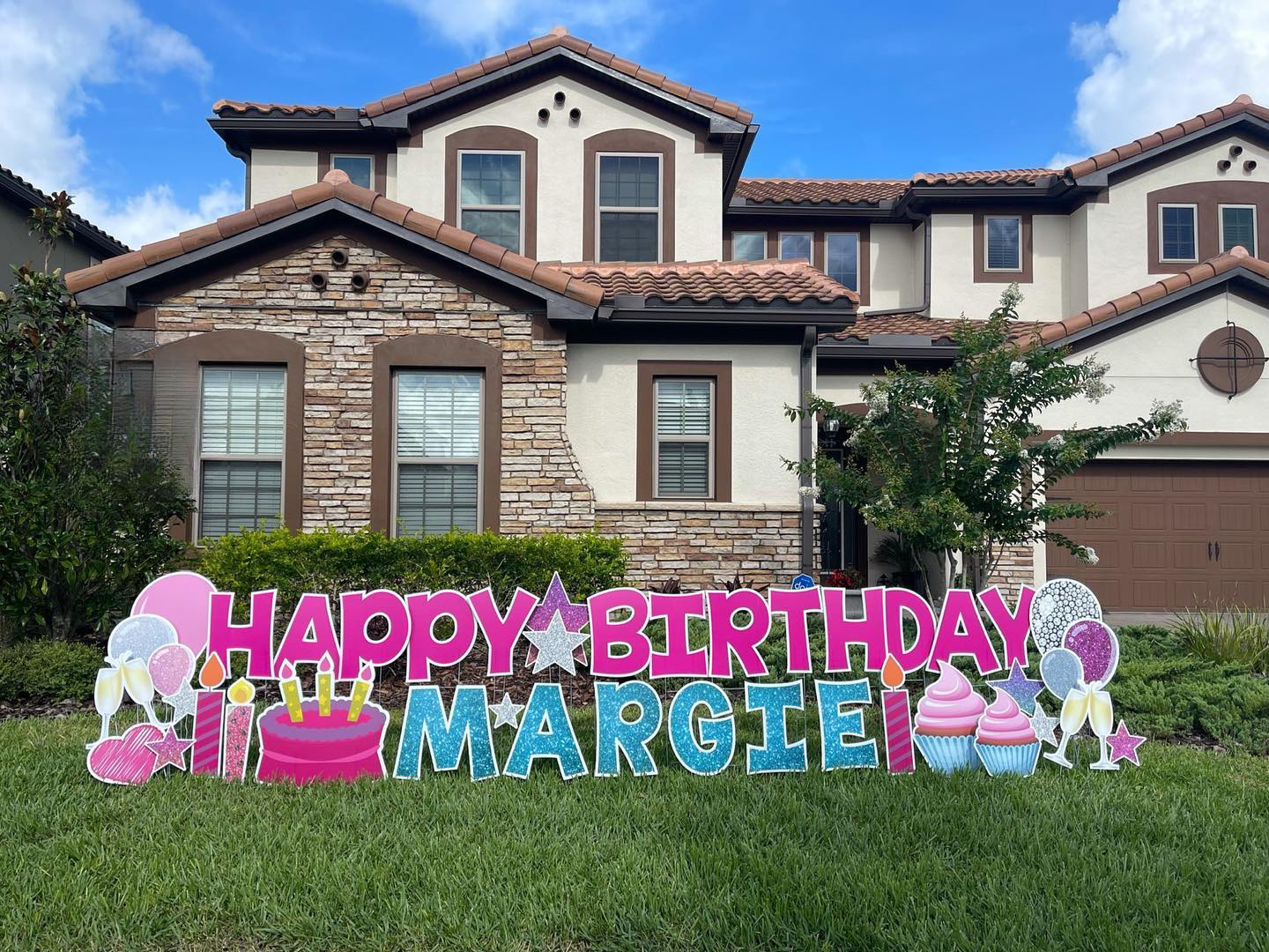 Colorful birthday yard sign in front of a house reads "Happy Birthday Margie" with images of candles, a birthday cake, cupcakes, balloons, and stars.